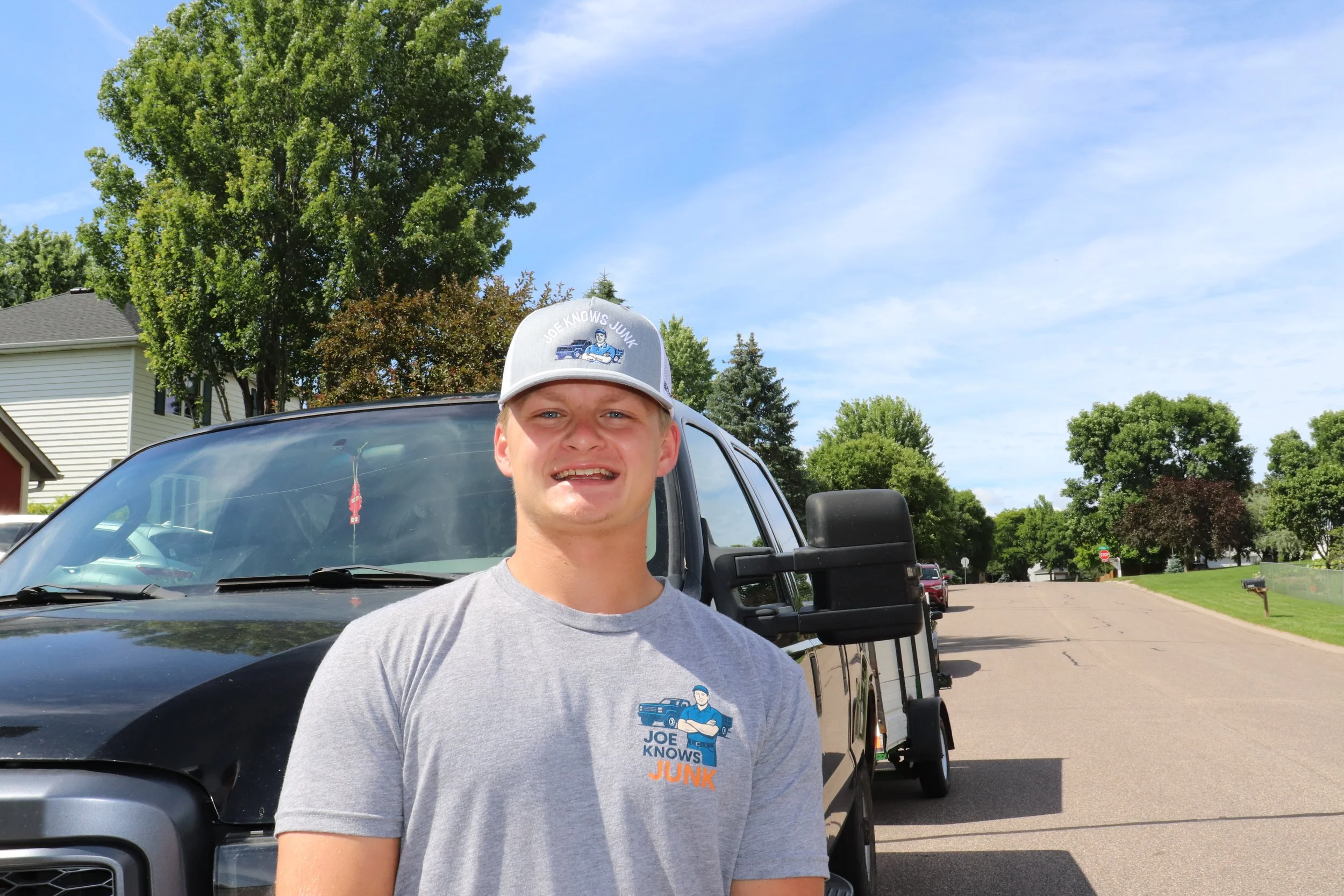 Young man outdoors smiling, wearing a gray T-shirt and a baseball cap, standing in front of a black pickup truck with a trailer attached, on a sunny day with clear blue sky, trees, and houses in the background.
