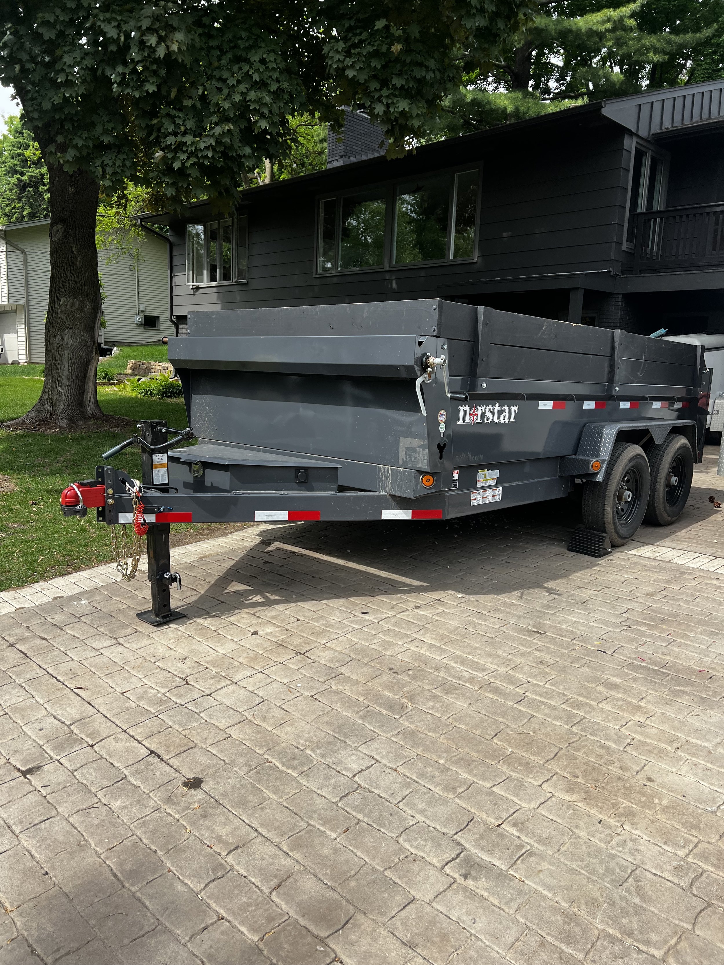A black utility trailer parked on a brick driveway in front of a black house with large windows and trees in the background.