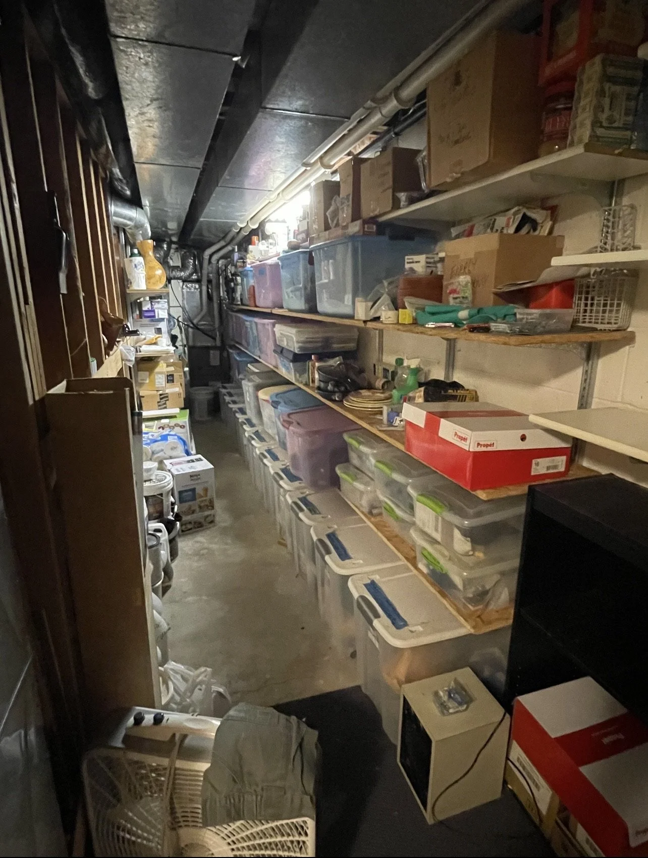 A narrow storage room with shelves filled with plastic bins, boxes, and various stored items, illuminated by overhead lighting and pipes running along the ceiling.
