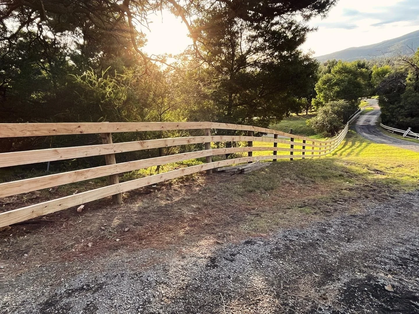 A rural dirt road with a wooden fence, trees, and mountains in the background, illuminated by the setting sun.