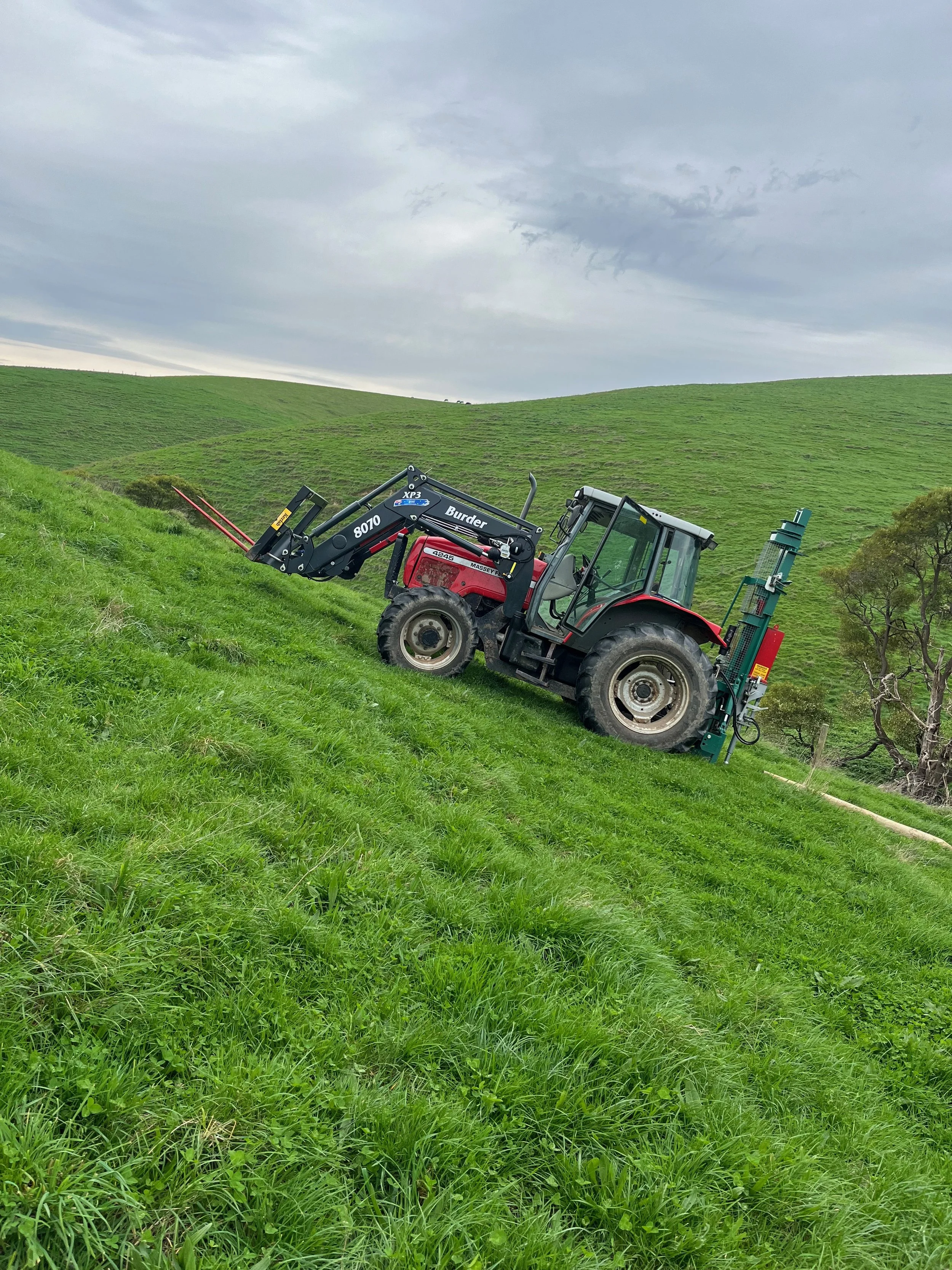 A red tractor with a black attachment on a sloped grassy hill, with green rolling hills and a cloudy sky in the background.