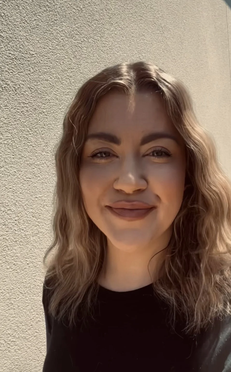 A woman with wavy, shoulder-length hair smiling outdoors against a textured beige wall.