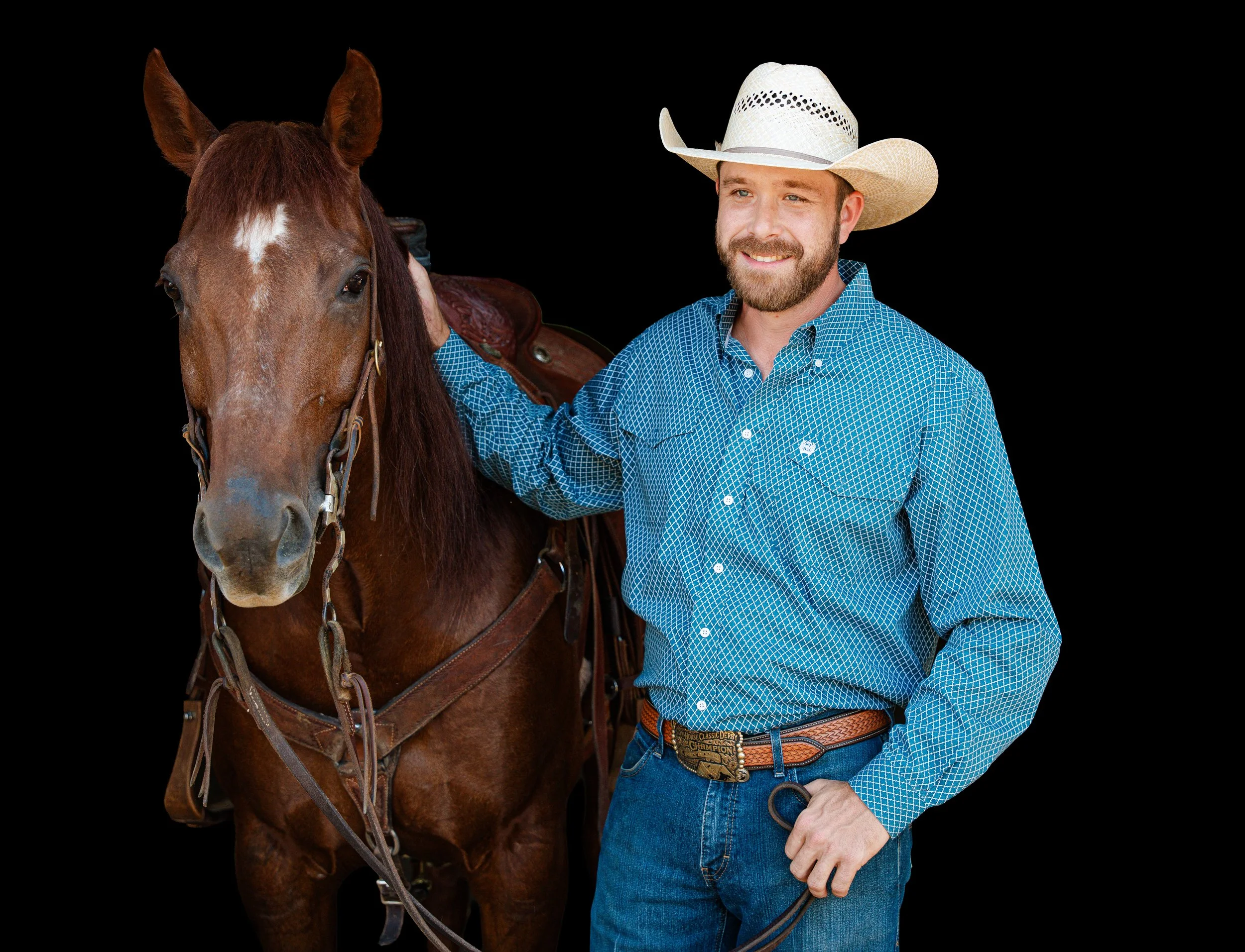 A man wearing a white cowboy hat, a blue checkered shirt, and jeans standing next to a chestnut rope horse with a saddle.