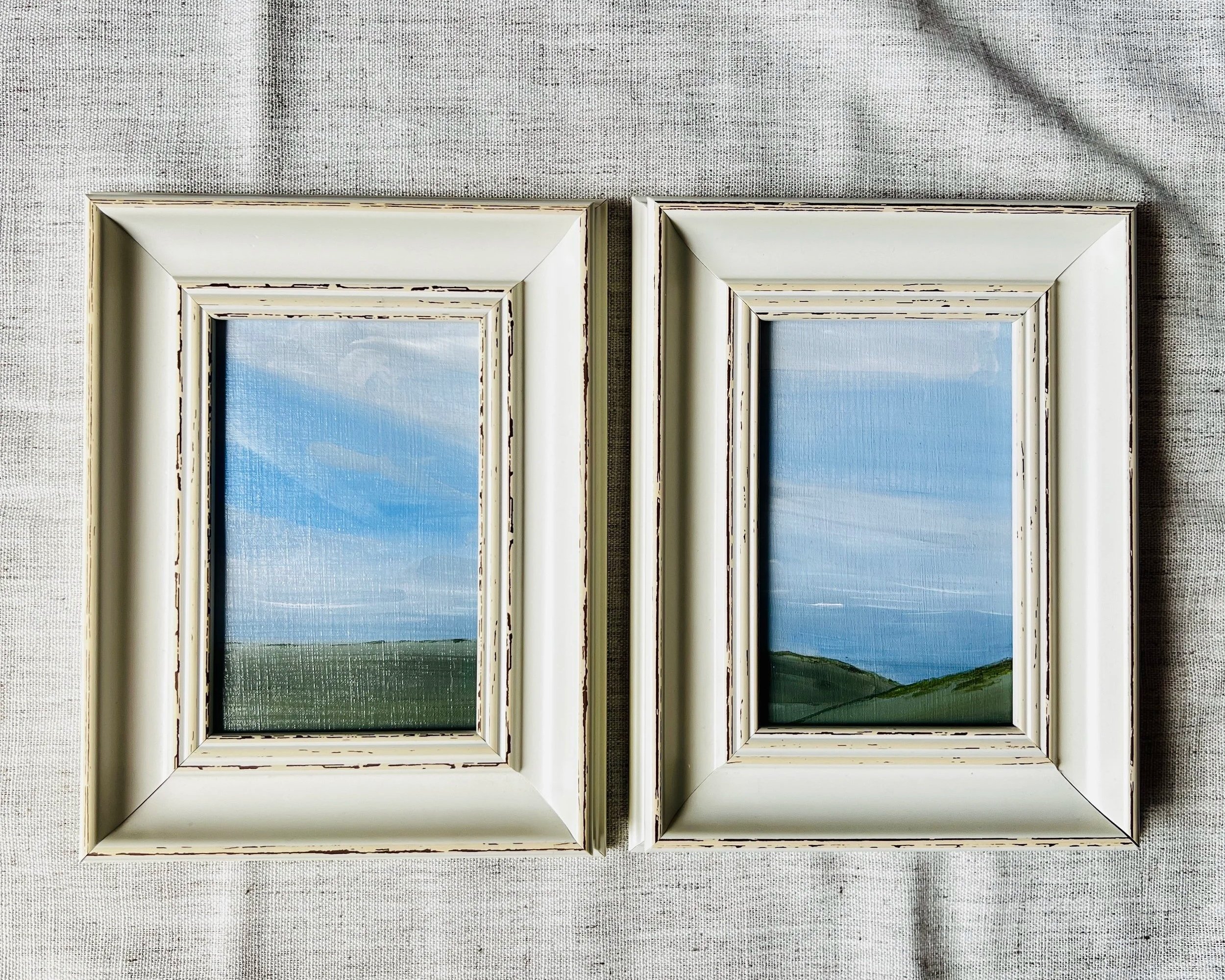 Two framed landscape paintings showing blue sky and green hills, placed side by side on a light-colored fabric surface.