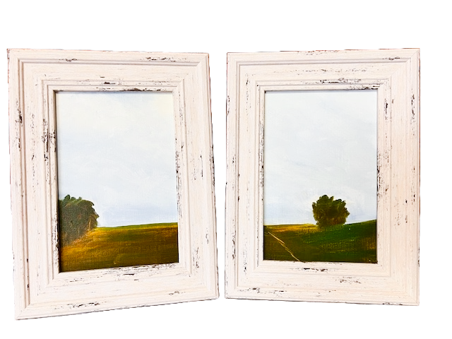 Two white wooden picture frames with landscape paintings of trees and open skies inside, leaning against each other.