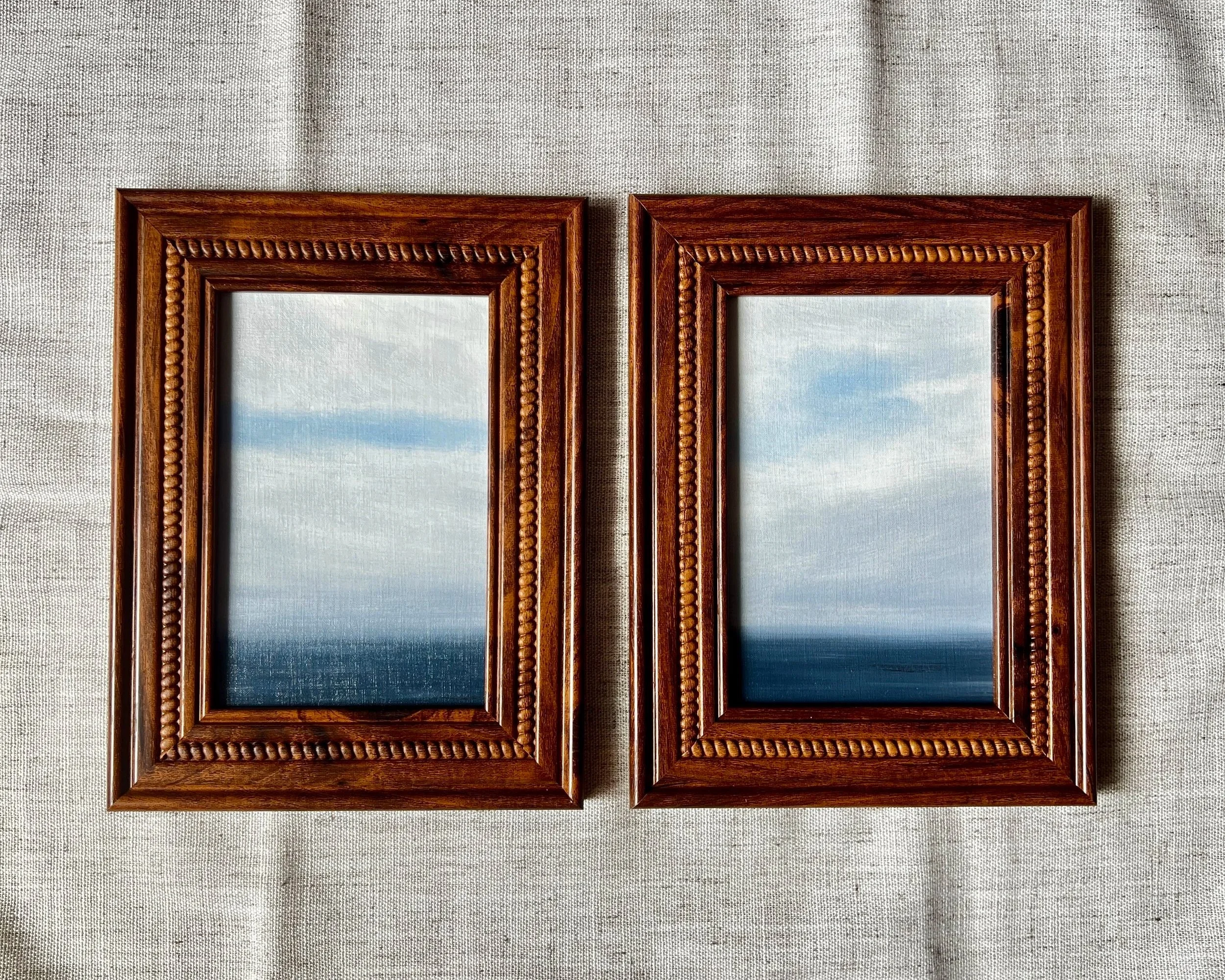 Two wooden picture frames hanging on a textured beige wall, each containing an image of a blue sky with clouds.