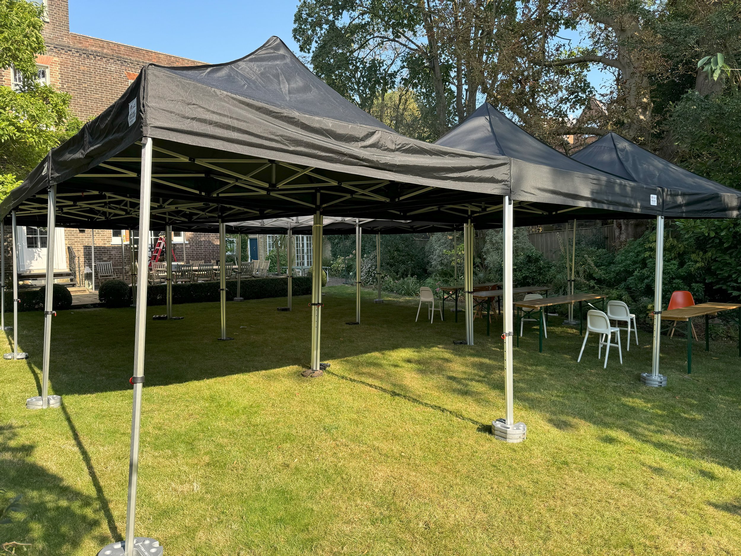 Large black canopy tents set up on a green lawn with tables and chairs underneath, surrounded by trees and a brick building in the background. marquee hire
gazebo hire
party tent hire
party marquee hire
event tent hire
wedding marquee hire
pop up gaz