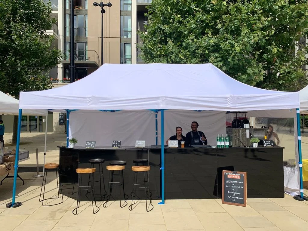 Outdoor bar setup under white canopy tent with three people behind the counter, set against an urban backdrop with trees and buildings.marquee hire
gazebo hire
party tent hire
party marquee hire
event tent hire
wedding marquee hire
pop up gazebo hire
