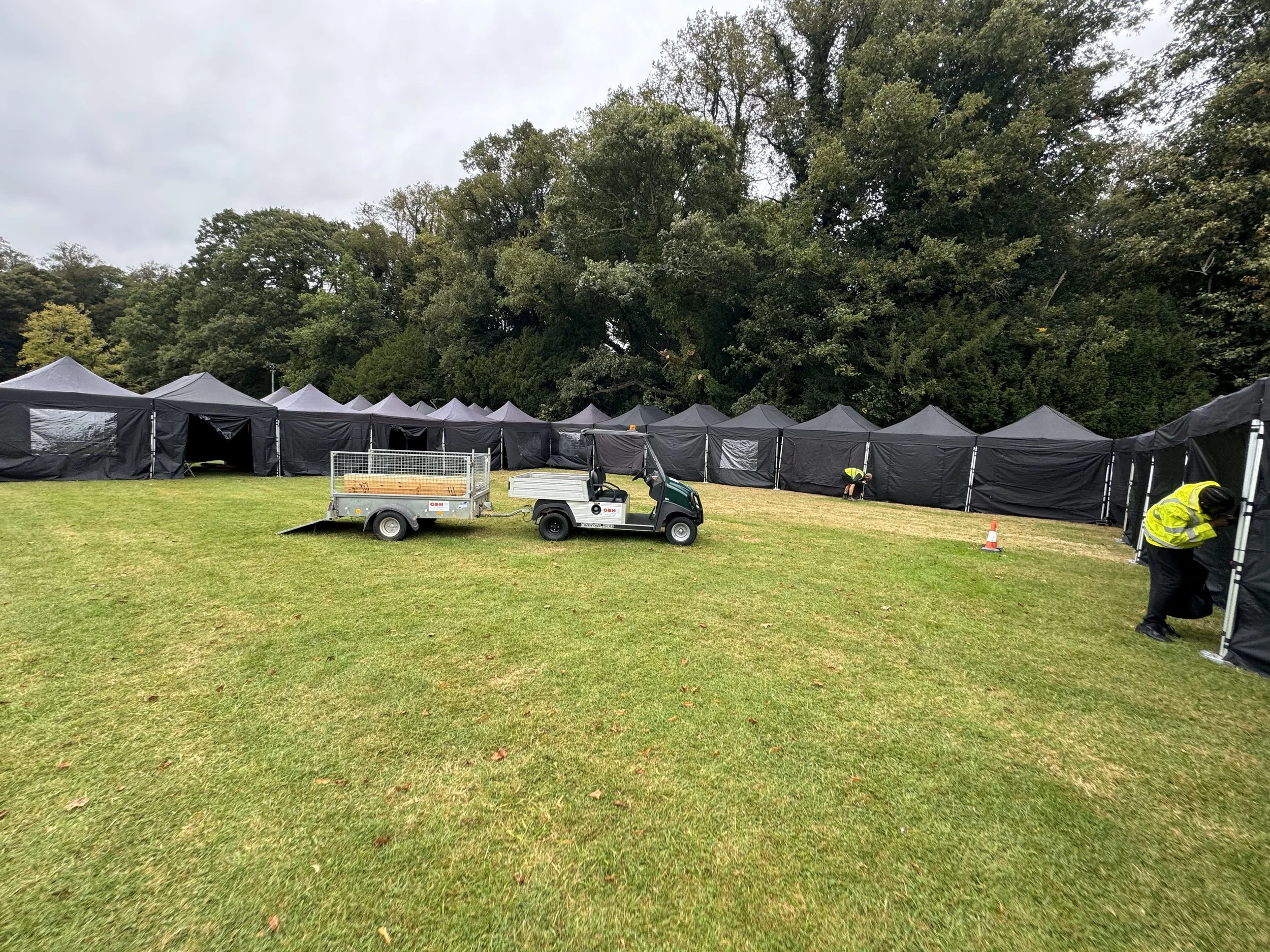 Black tents set up in a grassy outdoor area with trees in the background, and individuals working on the tent structures. marquee hire
gazebo hire
party tent hire
party marquee hire
event tent hire
wedding marquee hire
pop up gazebo hire
corporate ev