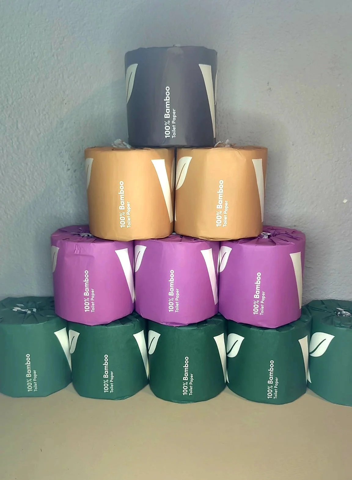 Stack of toilet paper rolls in black, brown, purple, and green packaging on a flat surface against a light-colored wall.