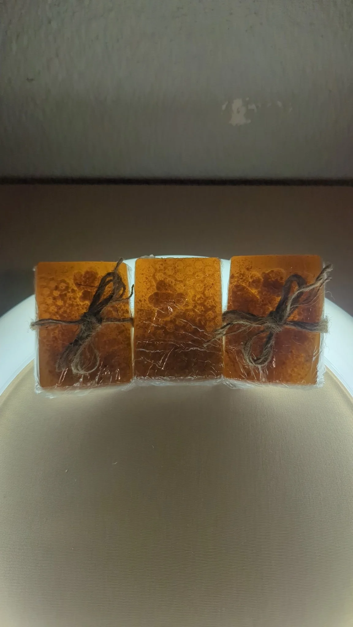 Three handmade soap bars tied with twine, wrapped in plastic, on a round surface with a grey wall in the background.