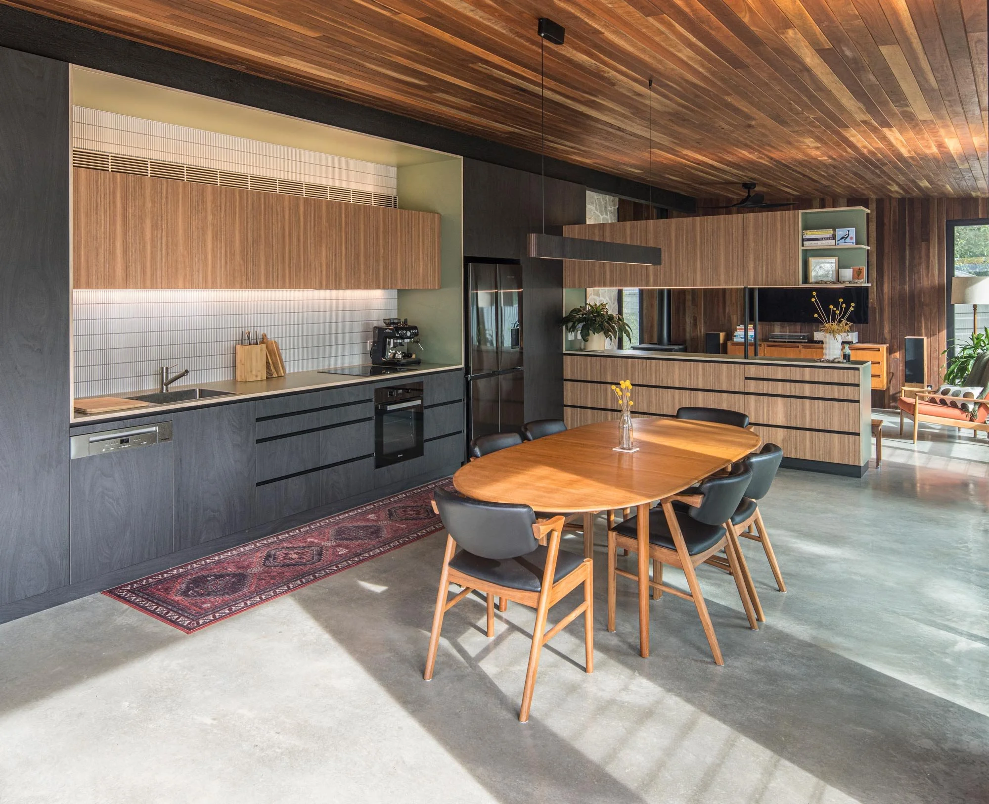 Angled view of the kitchen-dining area, highlighting layered materials: a polished concrete floor, red timber ceiling, and mid-century furniture. Black and brown kitchen cabinets with a green-trimmed, white-tiled inlet complete the style.