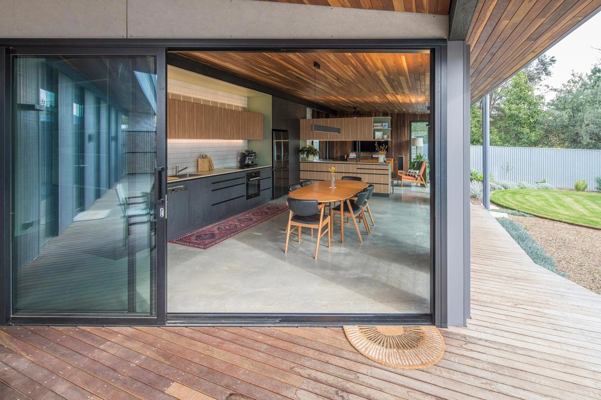 View through open terrace door into a kitchen-dining area with mid-century furniture and polished concrete floors. Black lower cabinets contrast with medium-brown upper cupboards, all tied together by a continuous red timber roof.