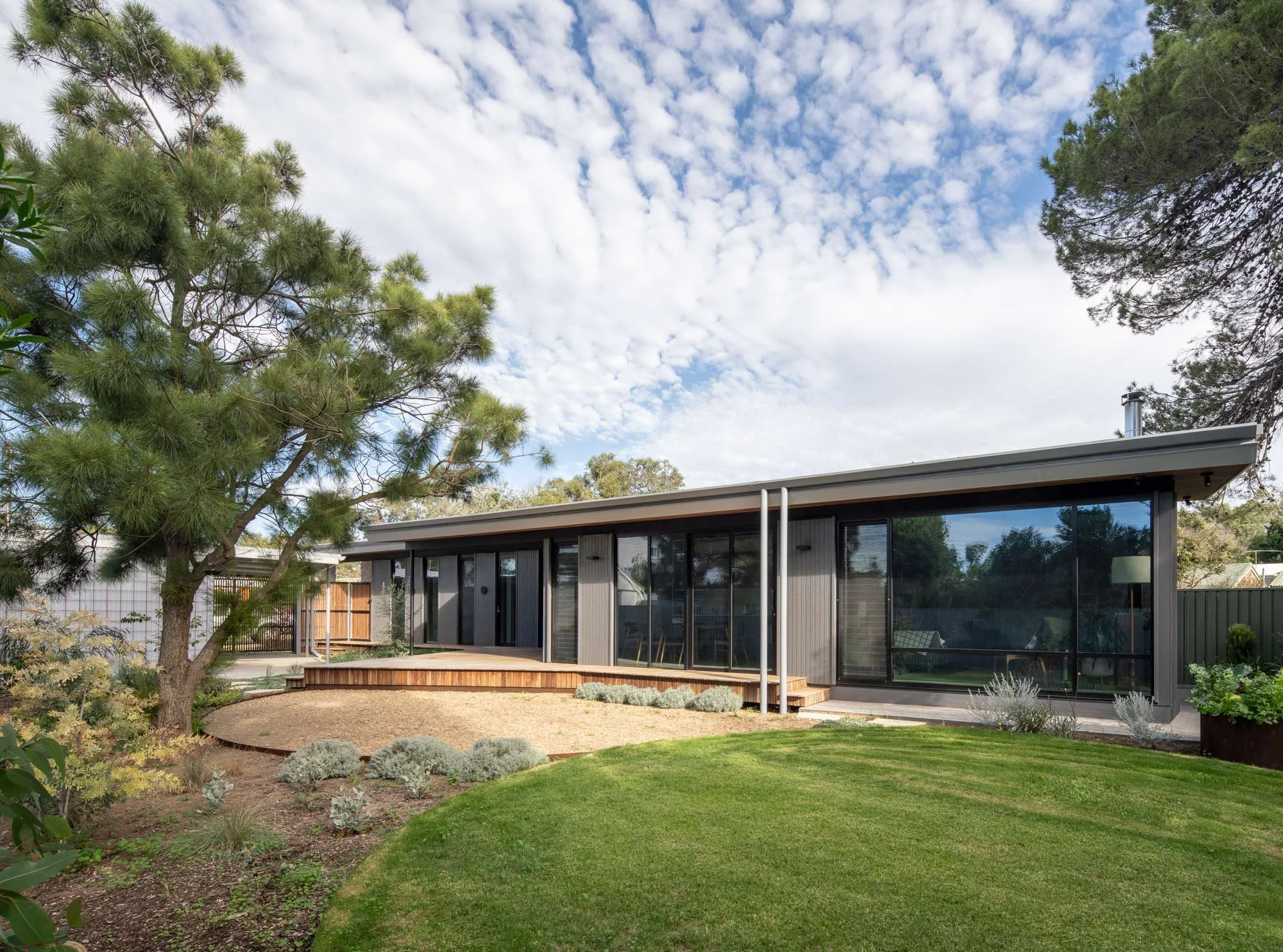 Side view of a flat-roofed bungalow from the garden. Large living and dining windows look out onto a tranquil landscape featuring a grassy lawn, a pebbled area, native shrubs, and a prominent pine tree.