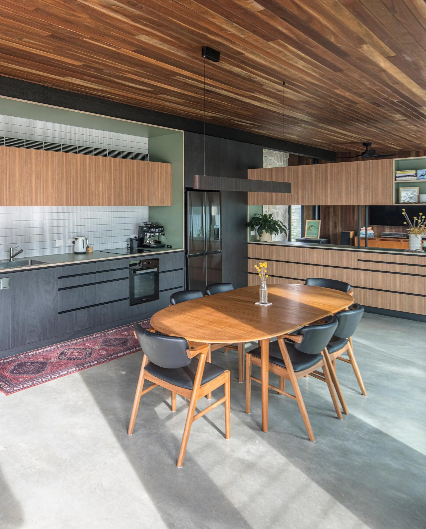 Angled view of the kitchen-dining area, highlighting layered materials: a polished concrete floor, red timber ceiling, and mid-century furniture. Black and brown kitchen cabinets with a green-trimmed, white-tiled inlet complete the style.