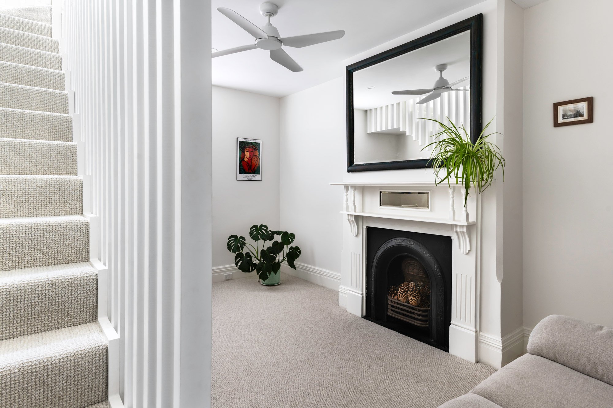 An all-white interior intentionally composed to show a modern, slatted wood staircase filling one-third of the frame, juxtaposed against a traditional Edwardian living room with an original fireplace filling the remaining two-thirds.