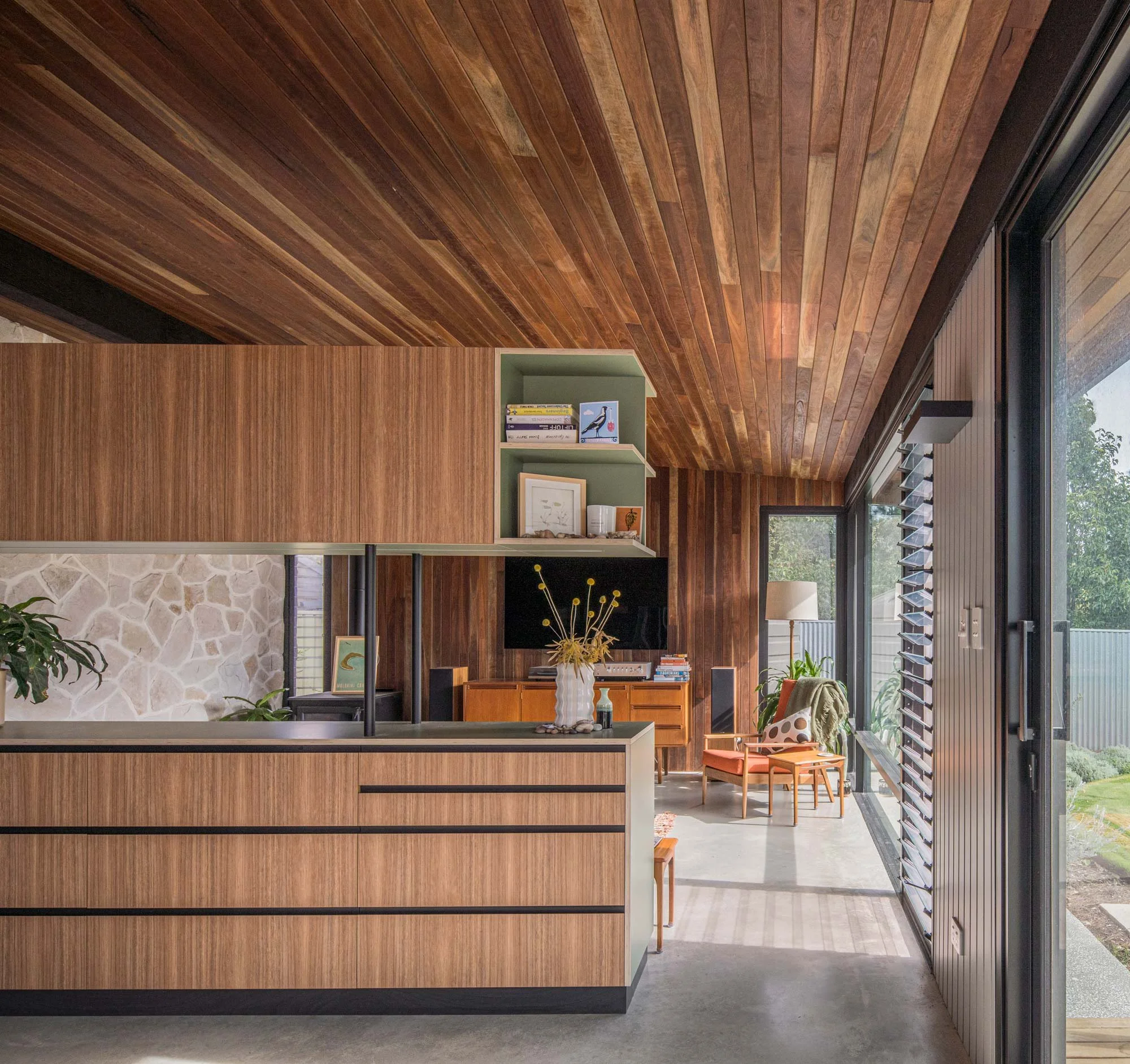 View from the kitchen into a mid-century living room. A wall clad in the same timber as the ceiling connects a garden-view window on one side to a texture-rich, crushed sandstone tile wall positioned directly opposite it.