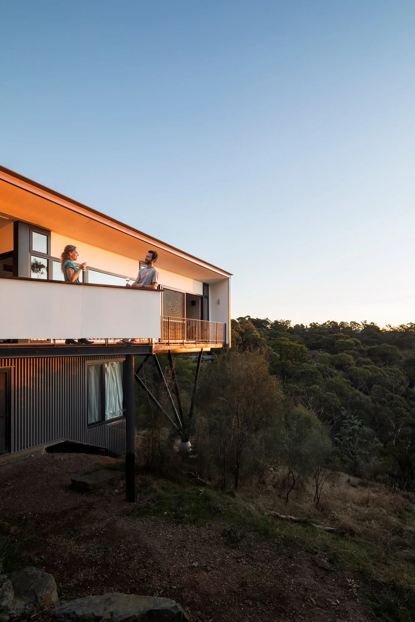 Angled view of two people drinking wine on a white-balustraded balcony of a modern stilt house, set against the Adelaide Hills bush.