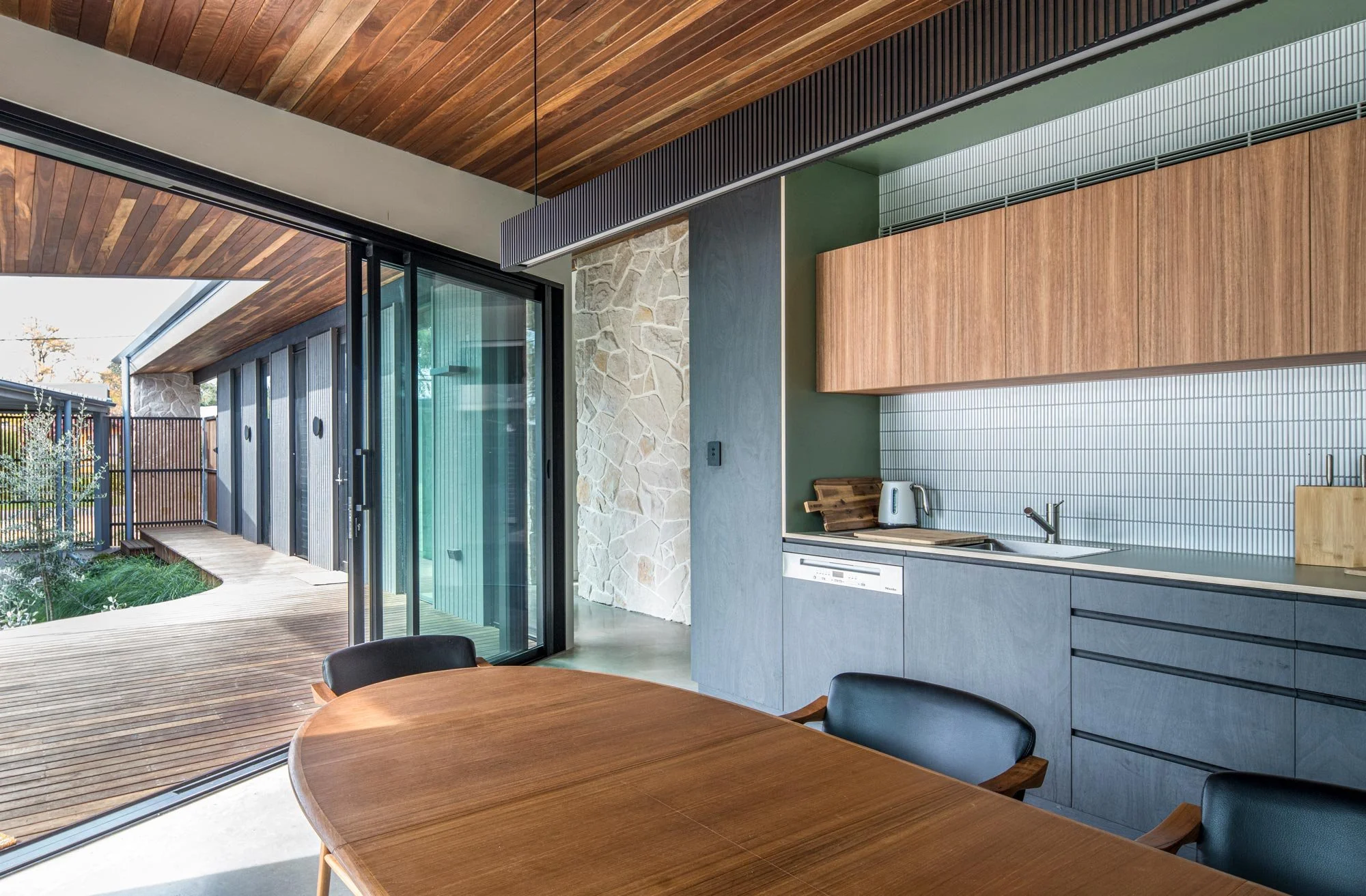 Mid-century kitchen-dining area opens to a deck with native landscaping. A red timber ceiling connects indoors to outdoors, highlighting black and brown cabinetry, polished concrete floors, and a sandstone-tiled hallway behind.