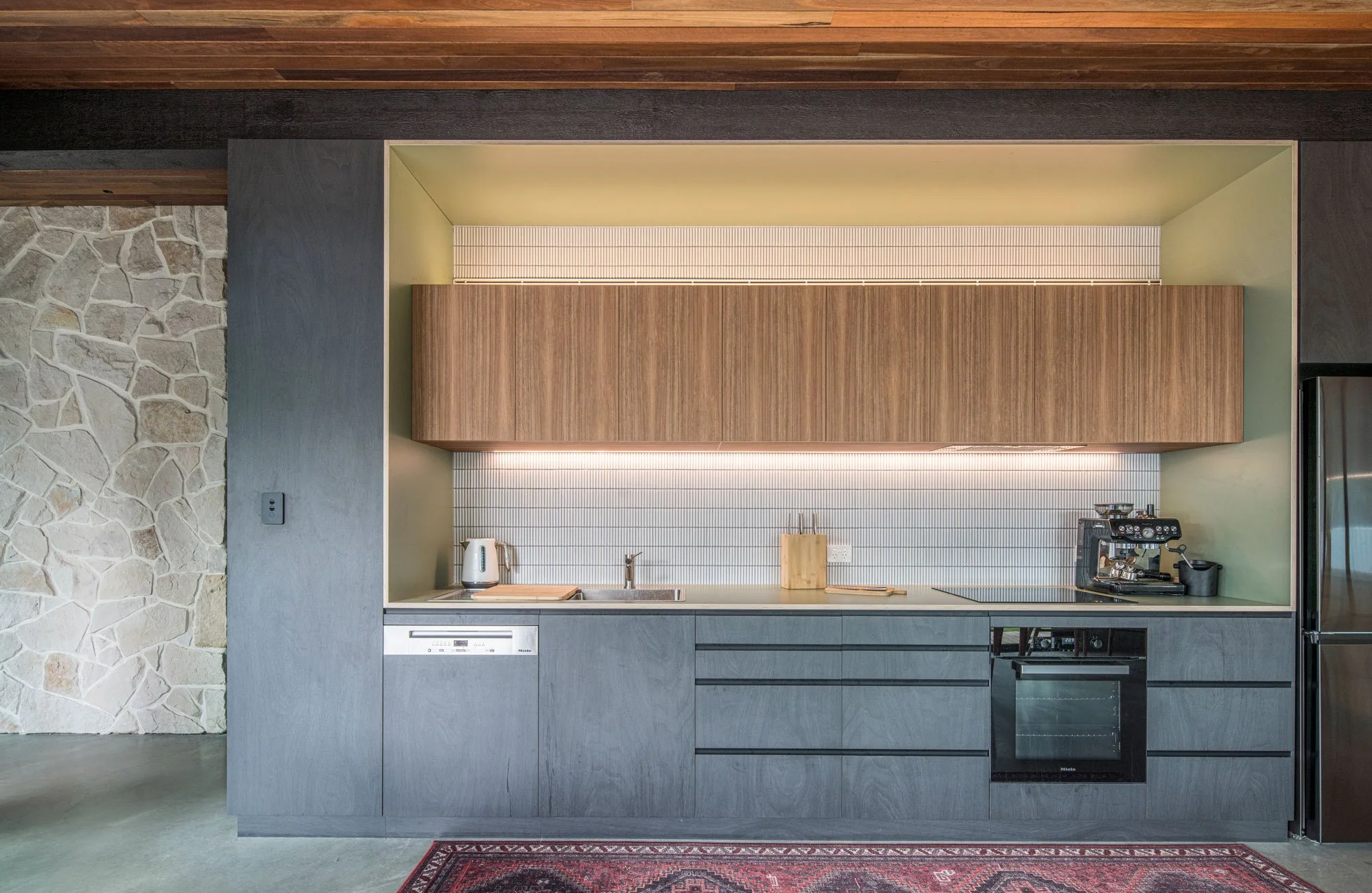 Front view of the kitchen, showcasing a layered mix of black, medium-brown, and green-trimmed timber cabinetry. This modern woodwork is placed directly alongside a feature wall of texture-rich, crushed sandstone tiles.