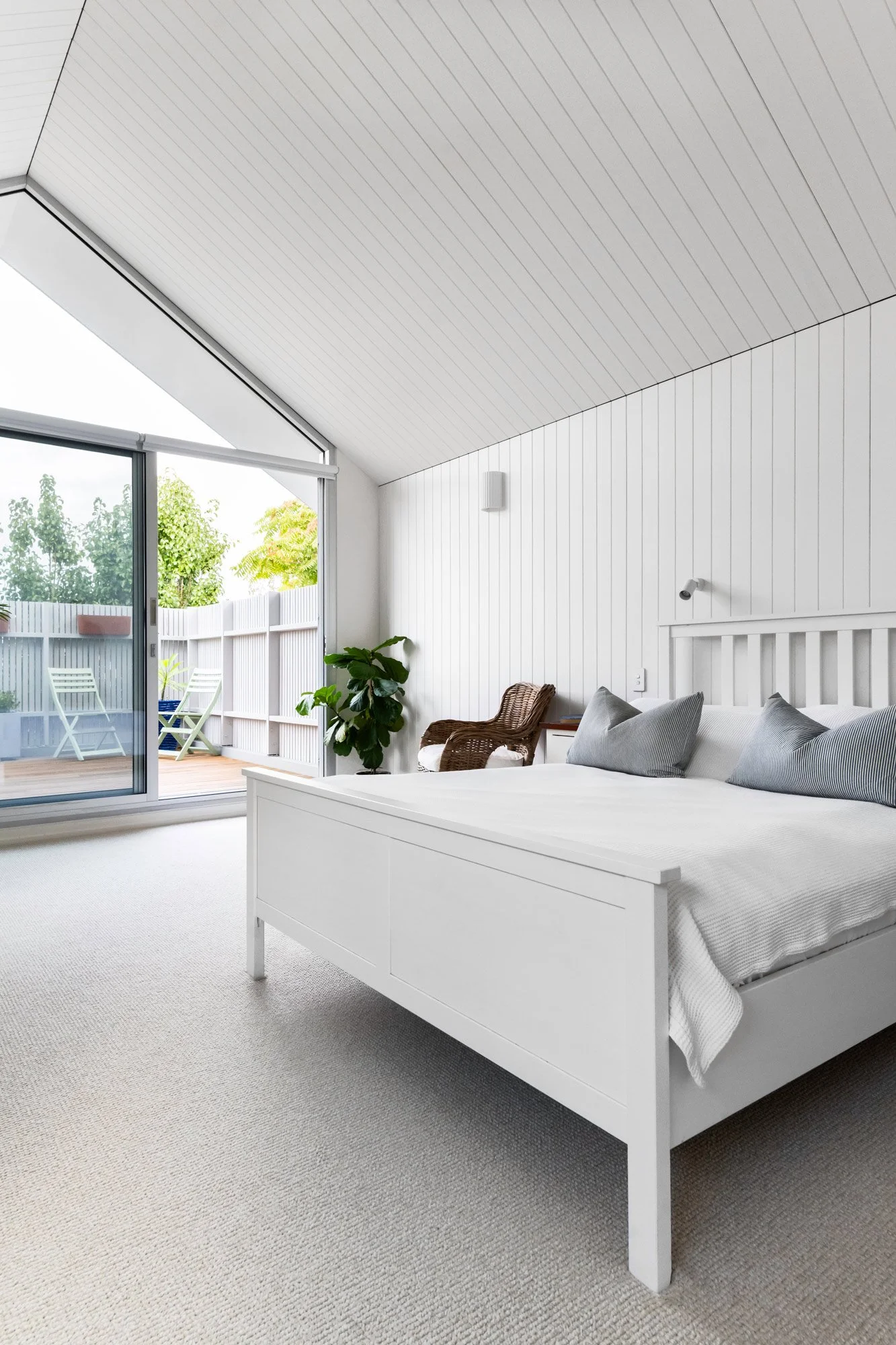 A low-angle view of an all-white bedroom, where wood boarding wraps seamlessly from the walls up into the high, gabled ceiling. A large window matching the roofline fills the room with light and leads to a terrace.
