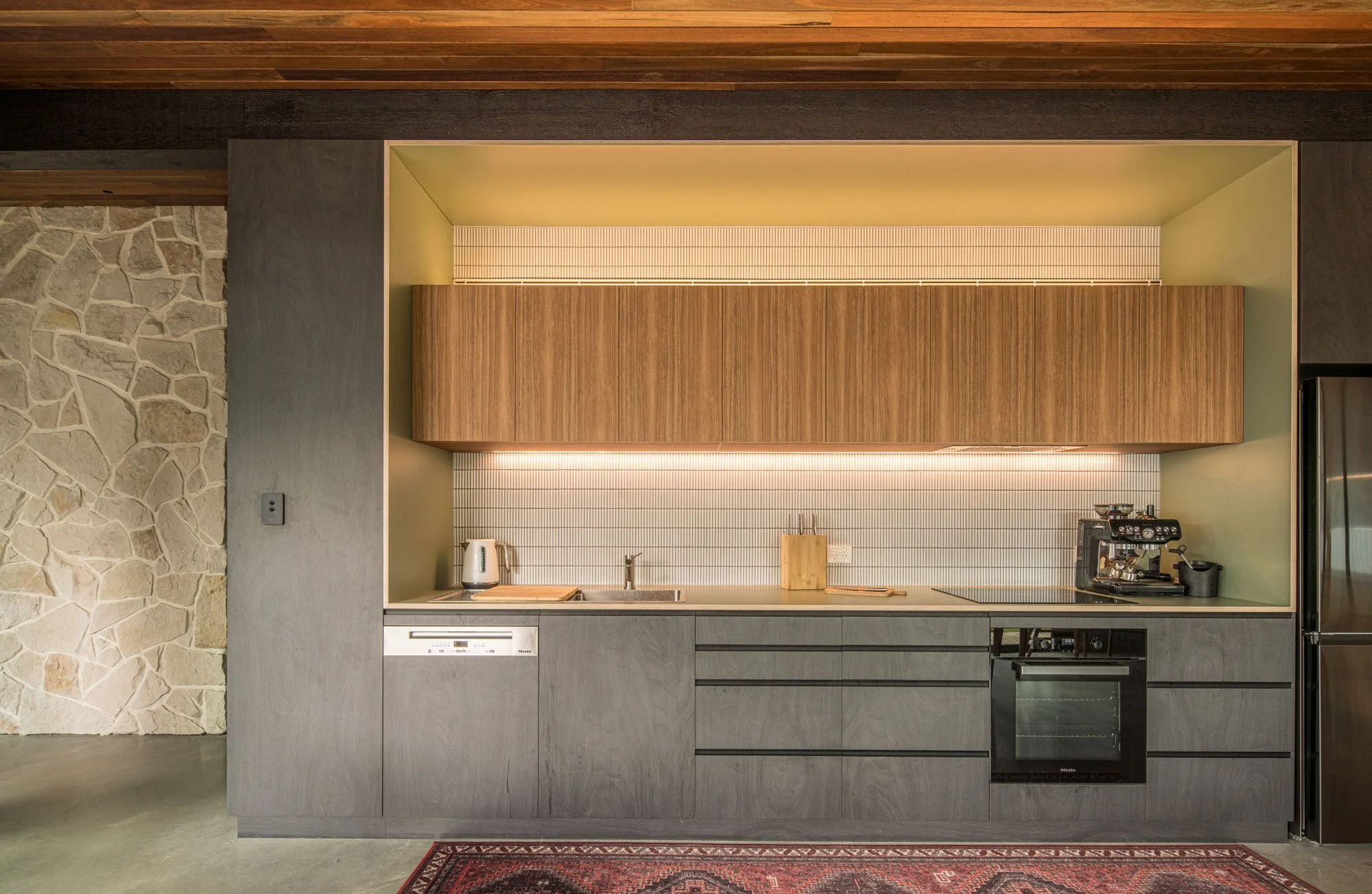 Front view of the kitchen, showcasing a layered mix of black, medium-brown, and green-trimmed timber cabinetry. This modern woodwork is placed directly alongside a feature wall of texture-rich, crushed sandstone tiles.