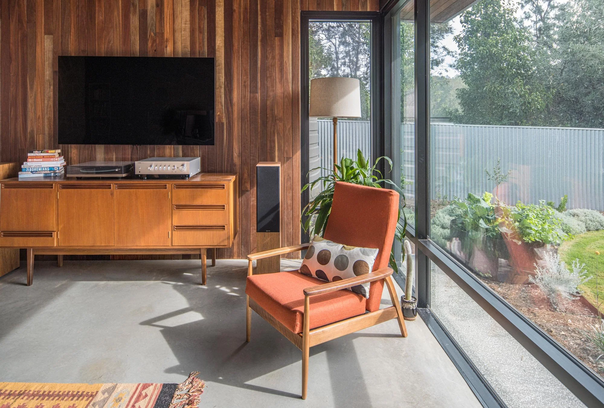 Corner view of a mid-century style living room, emphasising the rich timber wall panels and a large glass window that provides an expansive view over a native garden.