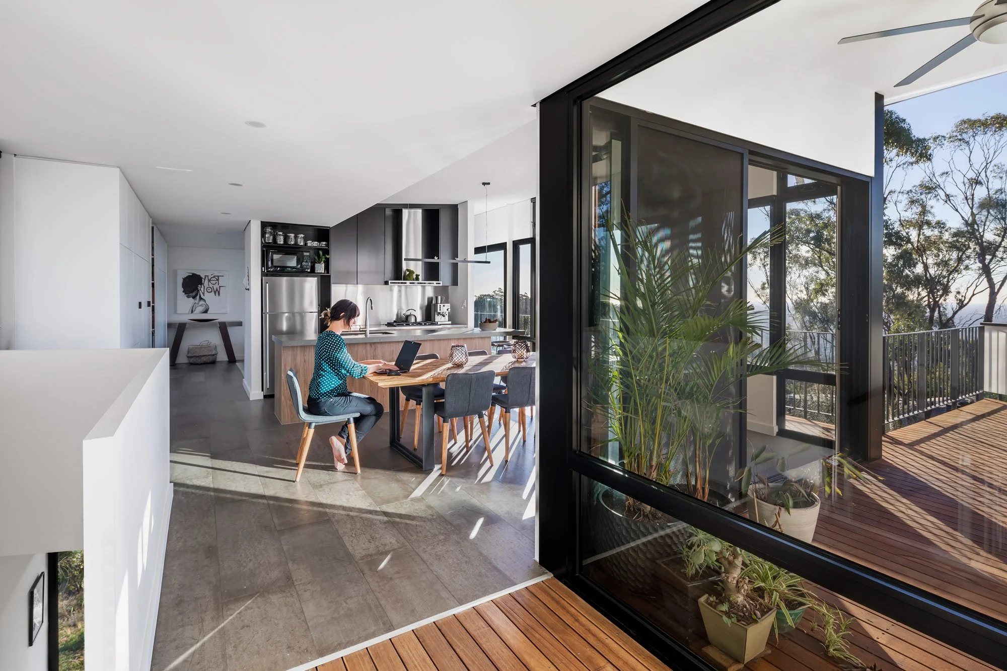 An angled view of a sun-flooded kitchen and dining area. To the left, a staircase leads downward, while a large window on the right offers a view of the wooden deck and the surrounding Australian bush.