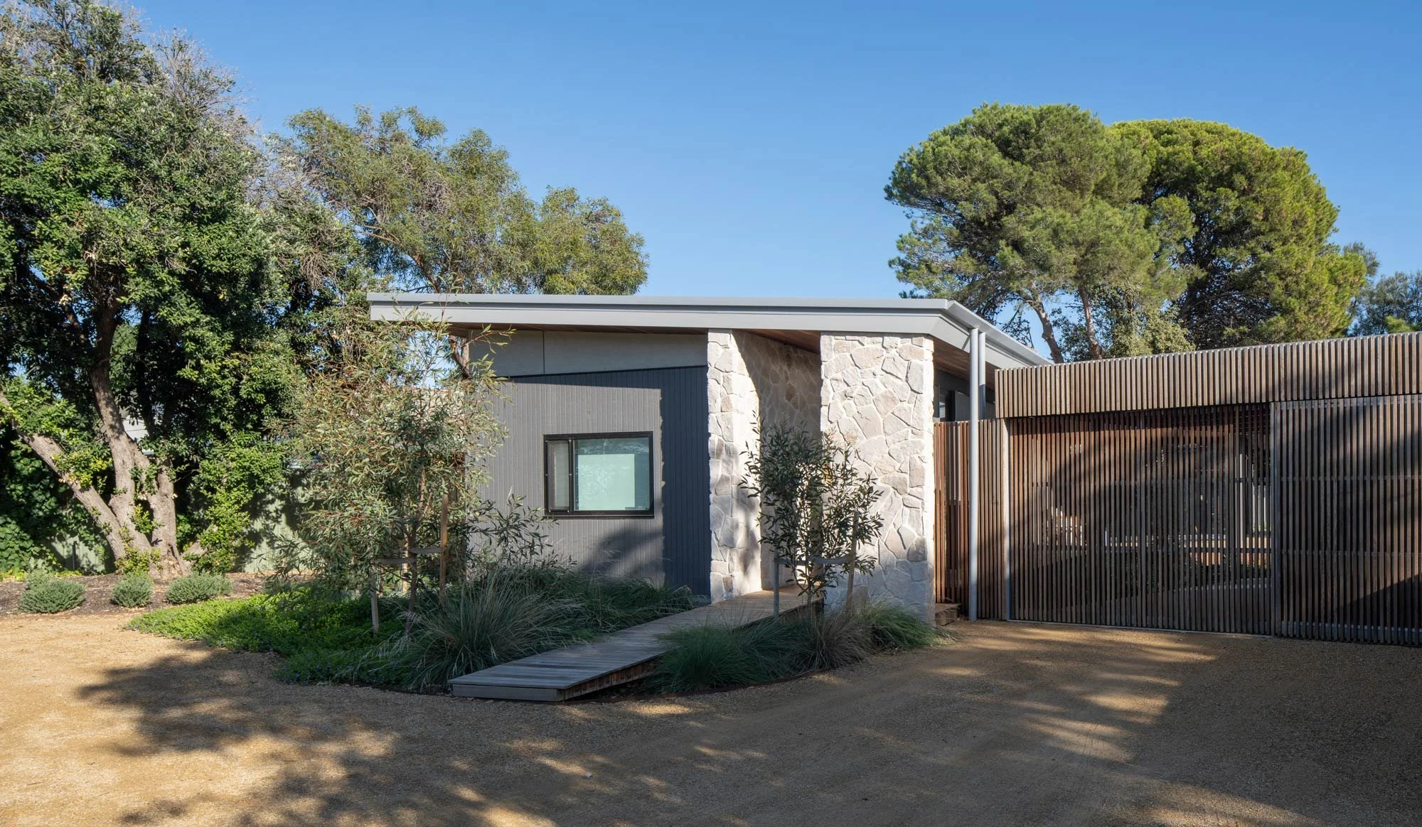 Flat-roof Palm Springs bungalow. A wooden footbridge framed by native Australian landscaping leads to a sandstone crushed-tile entrance. To the left is a grey cladded bedroom wall; to the right, a slatted natural wood garage.