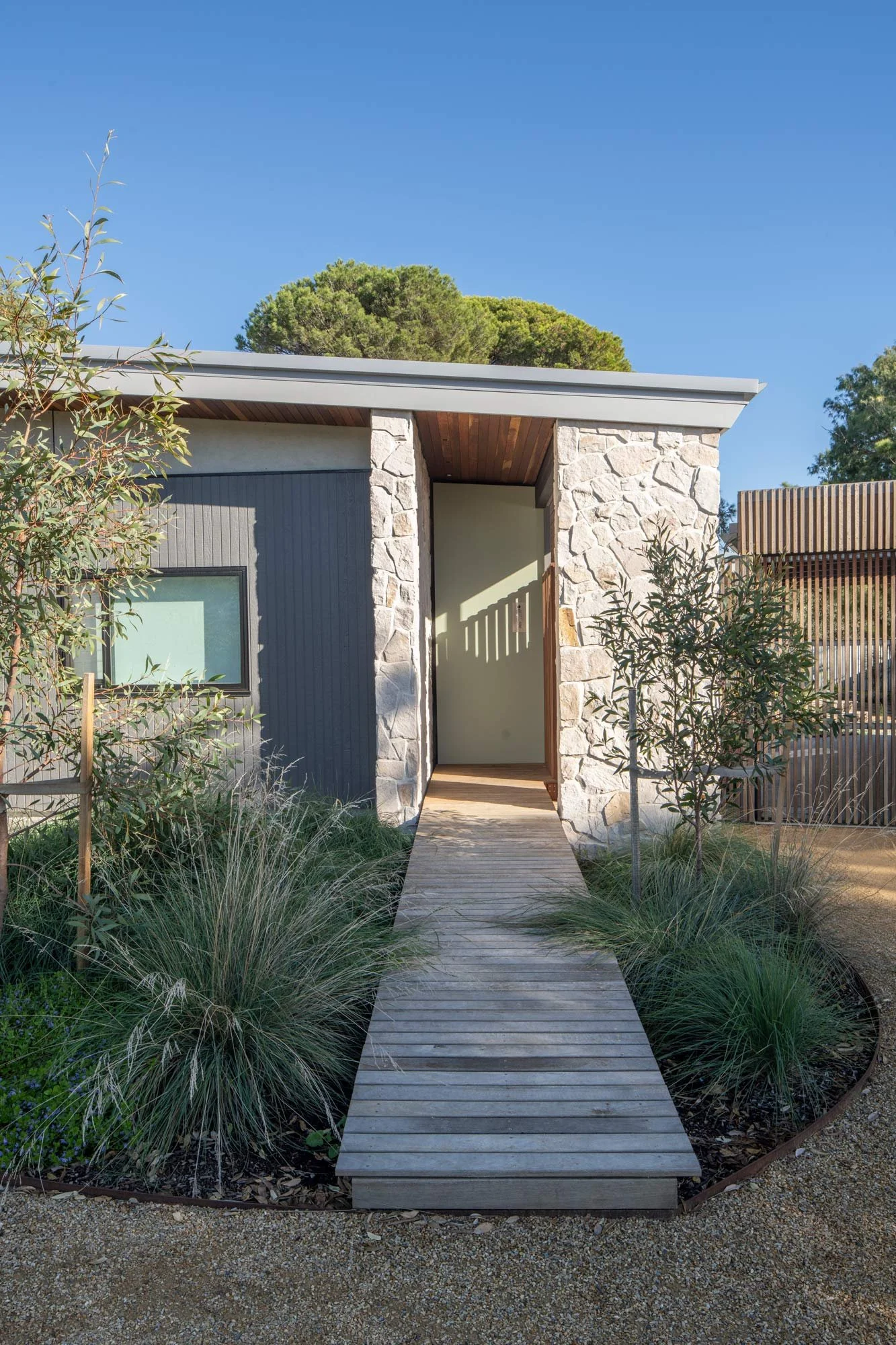 Front view of a bungalow entrance, where a wooden footbridge leads the eye to a sandstone crushed-tile entryway. Native Australian shrubs and eucalyptus trees frame the path, creating natural leading lines.