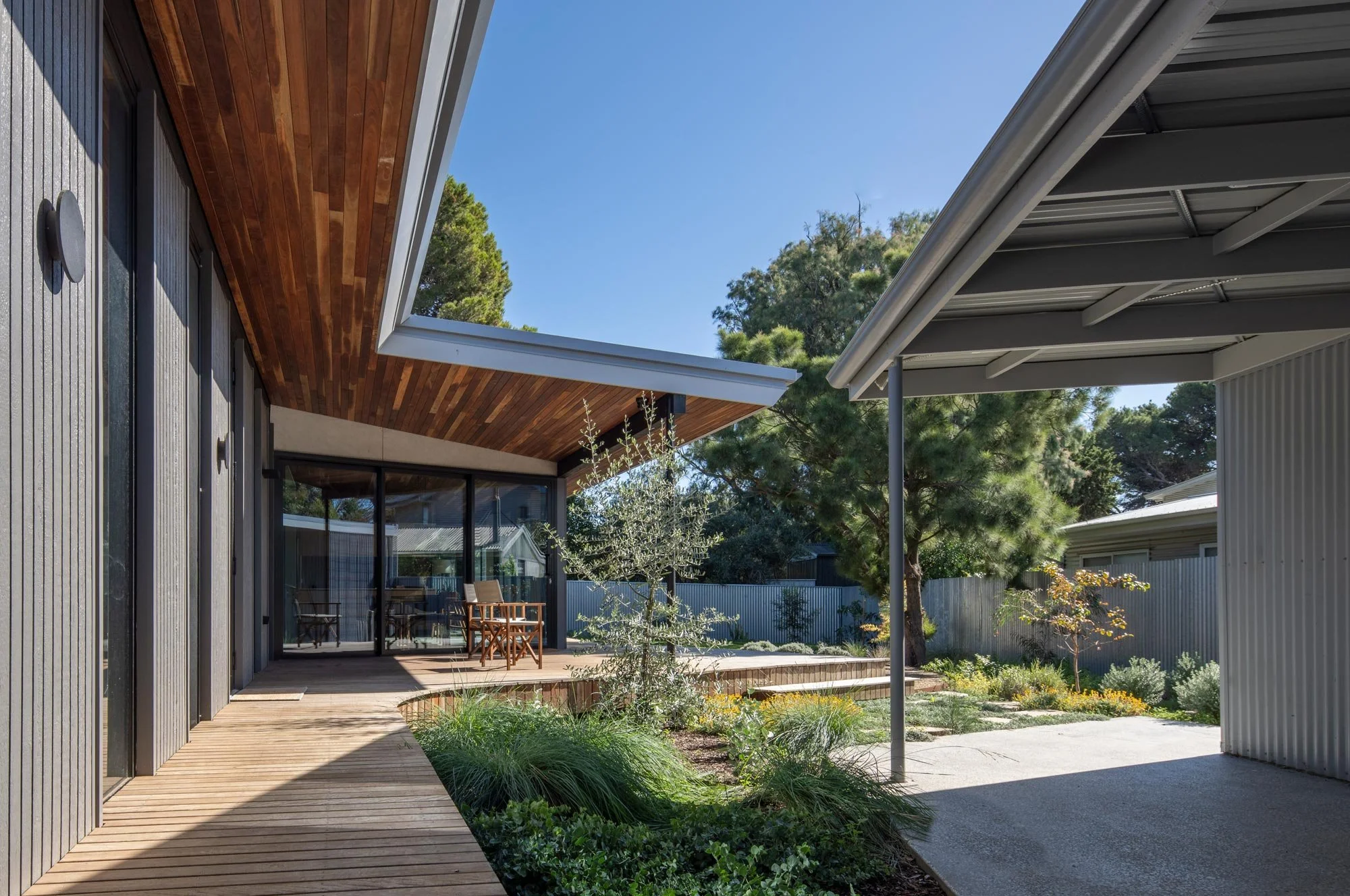 Low-angle view from a light brown timber deck, showing a contrast of materials. Features a grey wood-cladded house wall, a red timber under-roof ceiling, a large glass window, and a corrugated iron fence bordering the backyard.