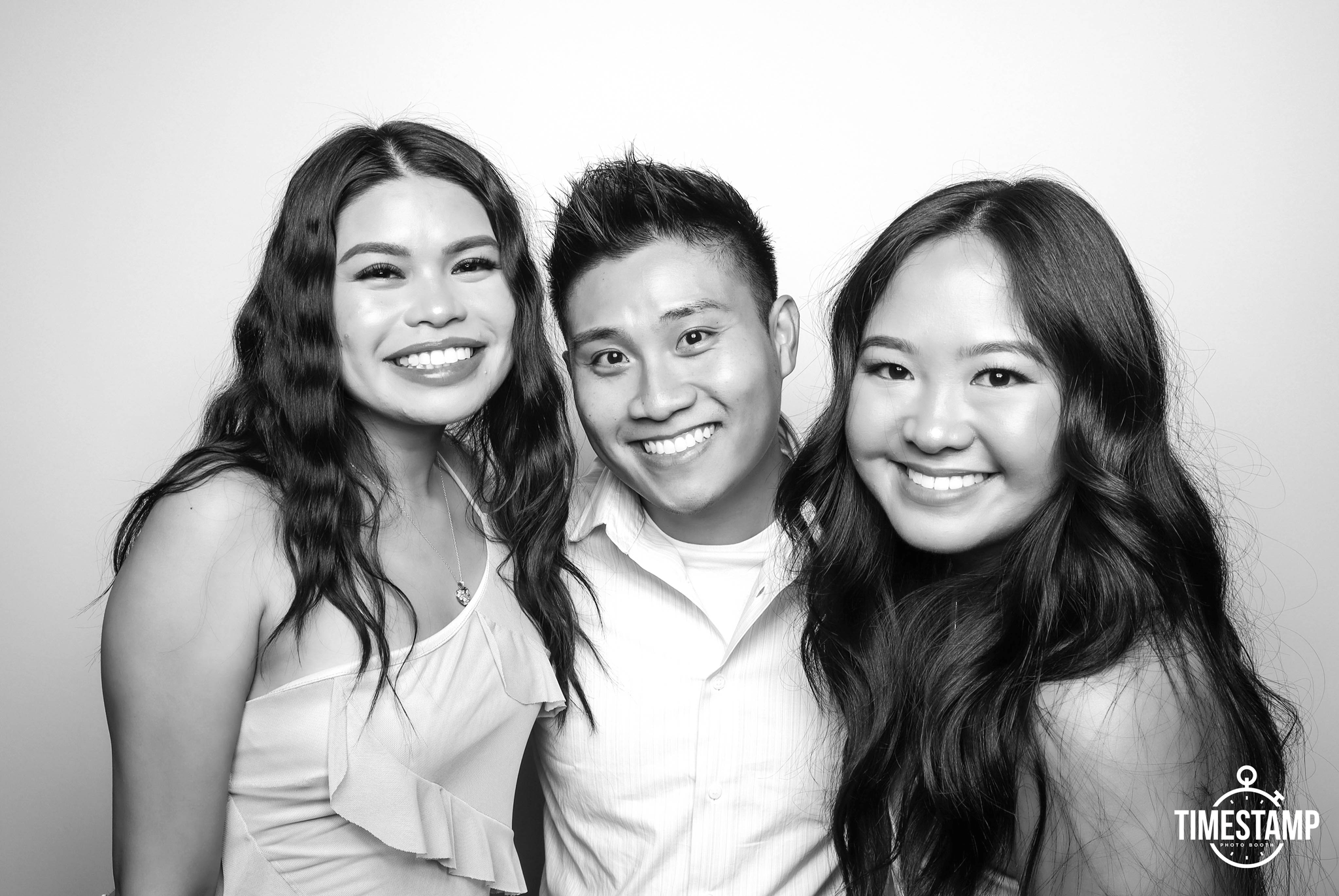 Black and white photo of three young smiling friends, two women and one man, posing closely together against a plain background.