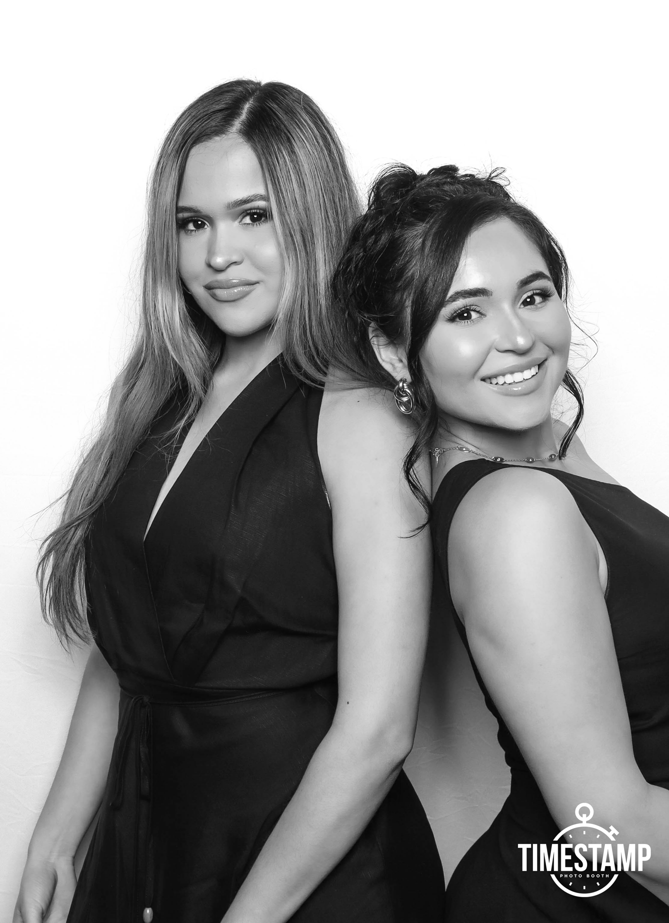 Two women wearing dark dresses, posing back-to-back and smiling in a photo booth.