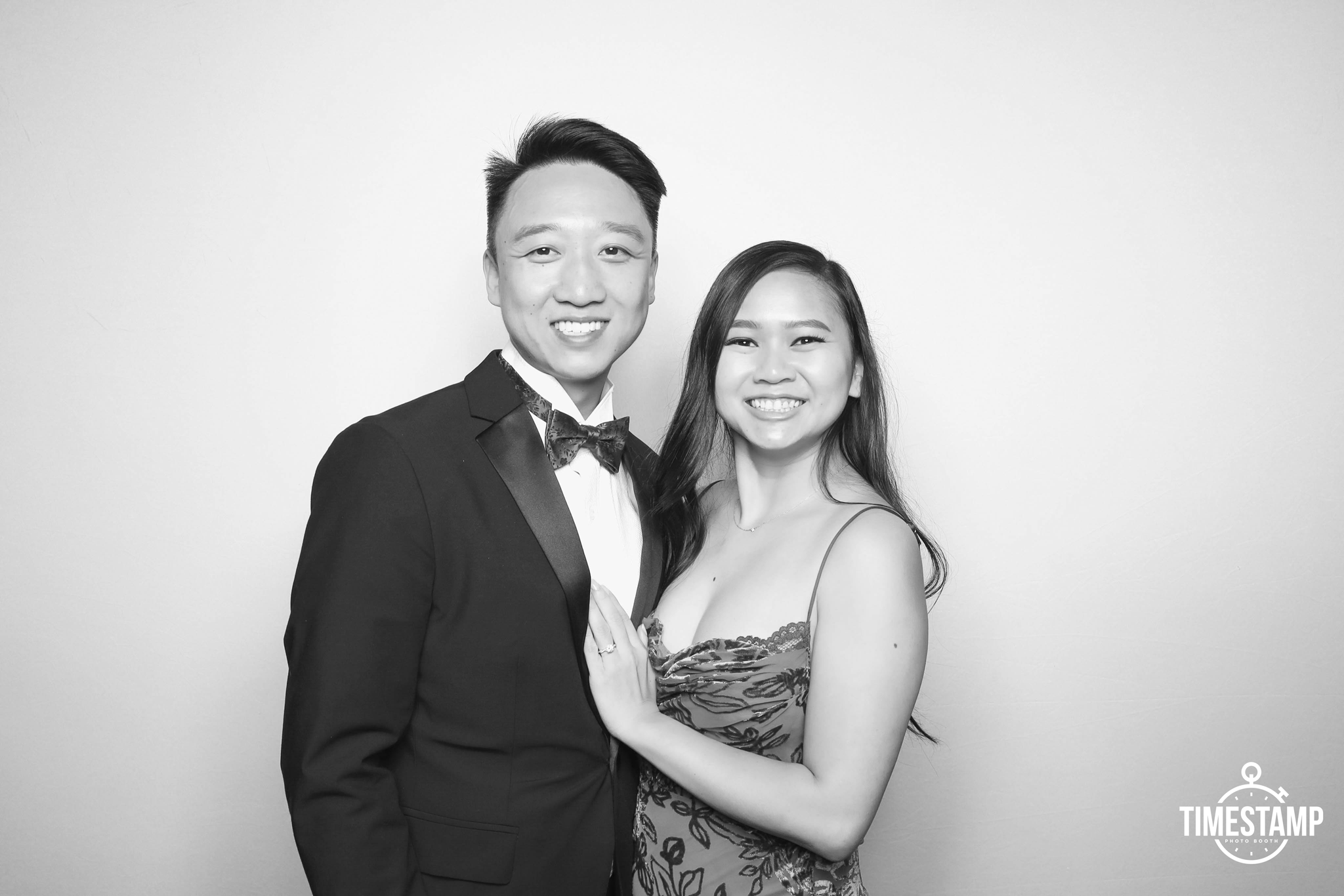 A young couple dressed in formal attire posing together, smiling at the camera, against a plain background. The man is wearing a tuxedo with a bow tie, and the woman is wearing a spaghetti strap dress with floral pattern.
