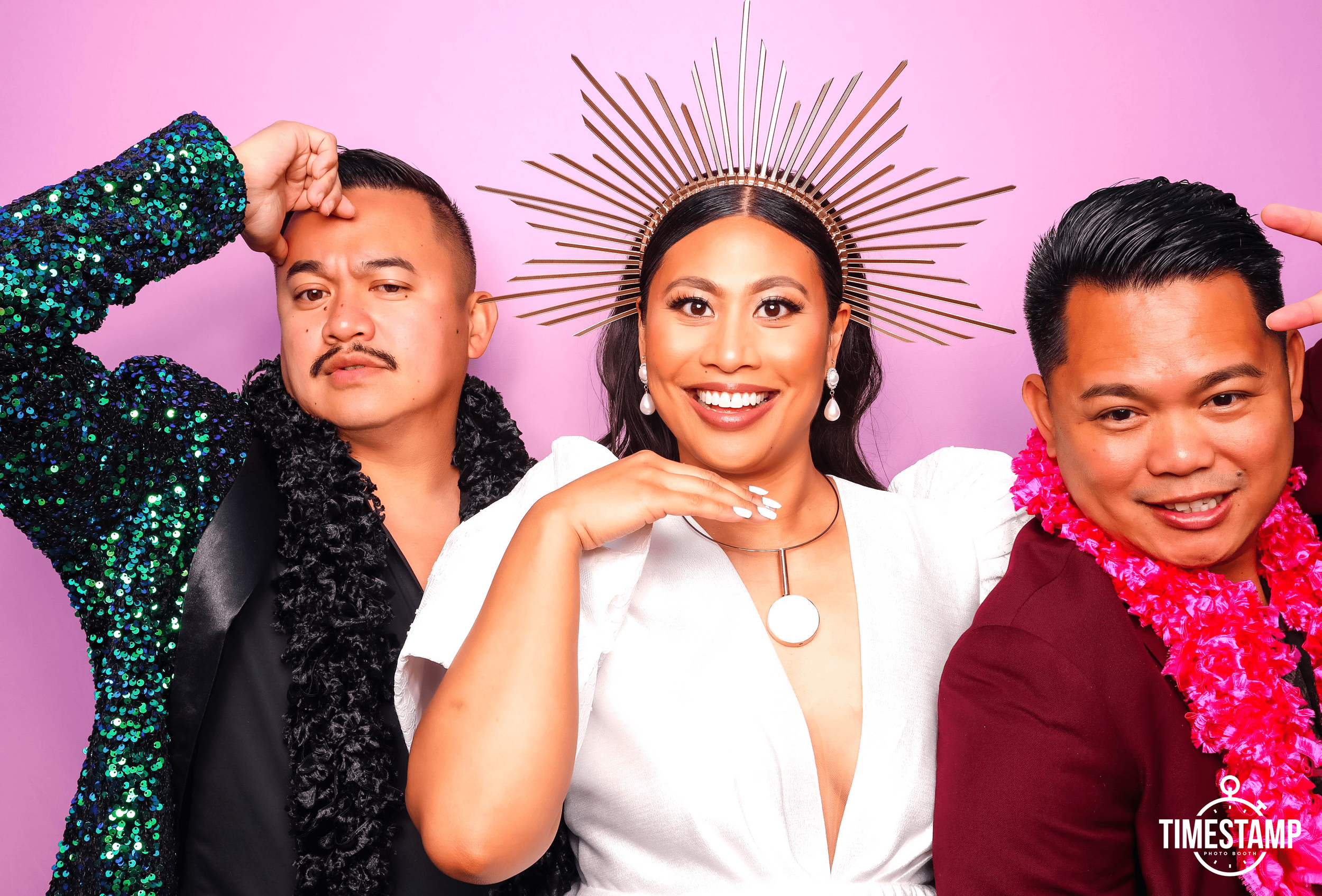 Three people posing for a photo at a photo booth. The woman in the middle is smiling, wearing a white dress, a large halo-shaped headpiece, and jewelry. The man on the left is wearing a dark outfit with a shiny, sequin jacket and has a half-smile with a hand raised to his head. The man on the right is smiling, wearing a dark red jacket, and has a pink lei around his neck. The background is pastel pink.