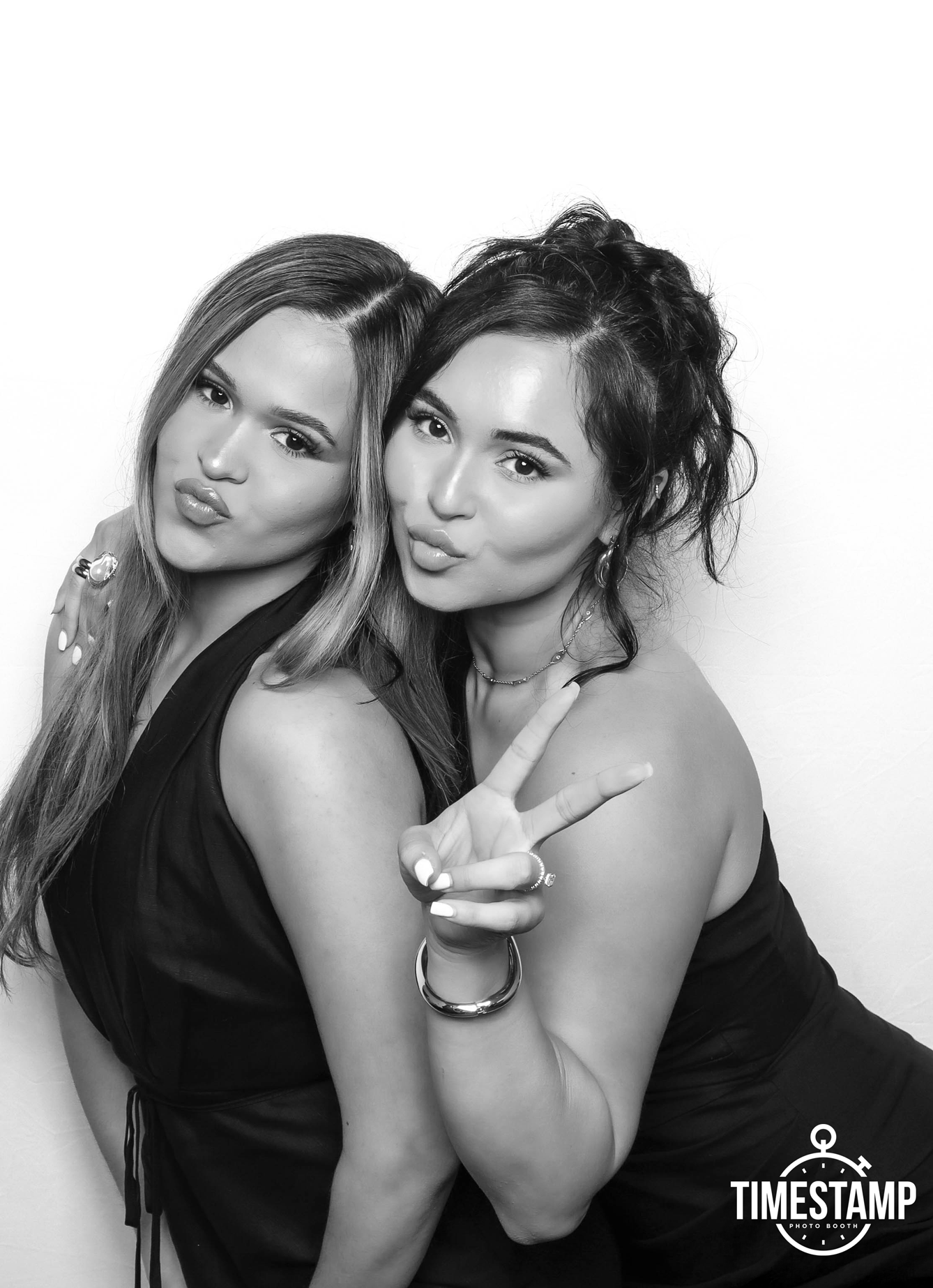Two women posing together in a photo booth, one making a kiss face and the other flashing a peace sign, against a plain background, with the TIMESTAMP logo in the bottom right corner.