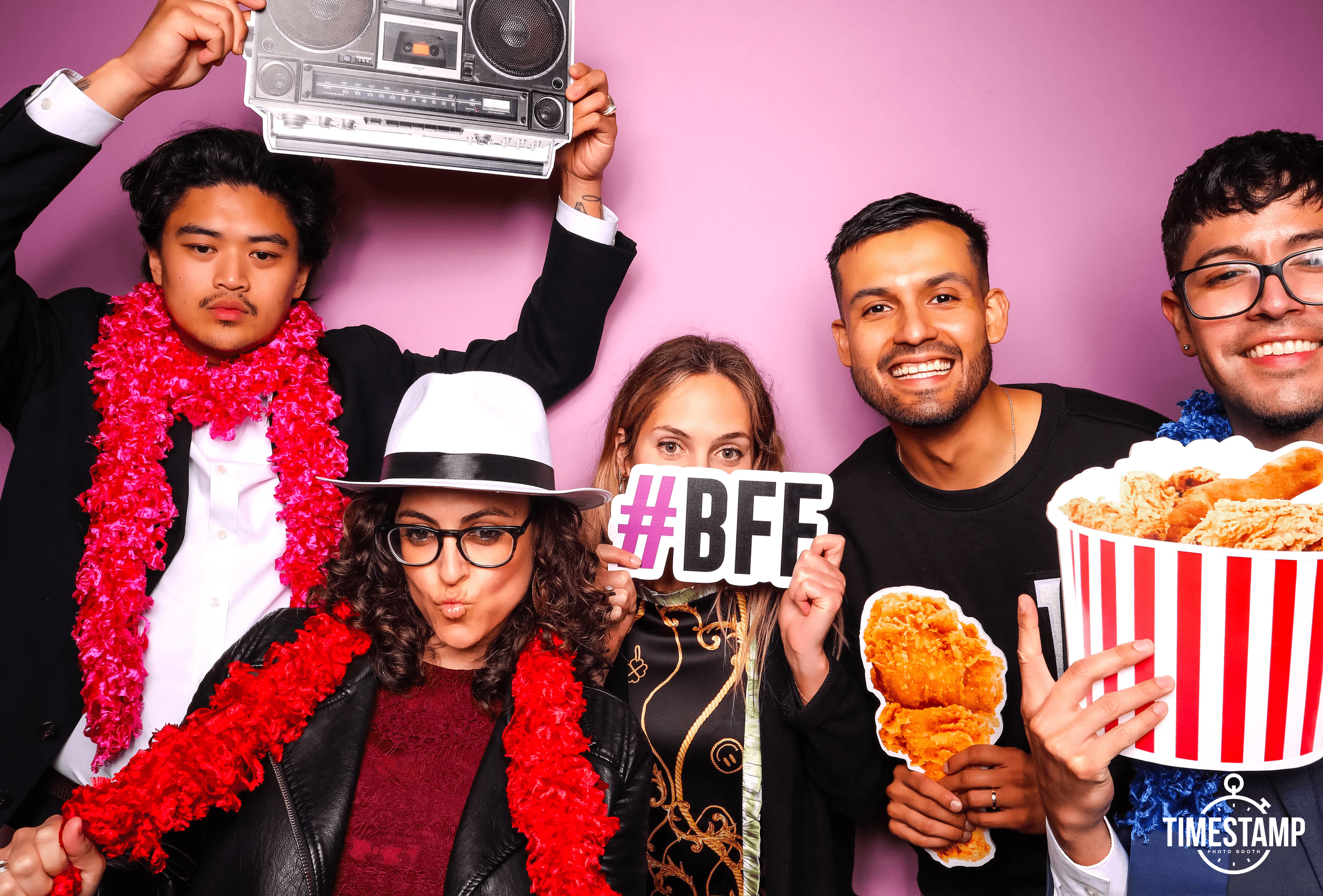 Group of five friends celebrating in photo booth with props, holding a '#BFF' sign, a large bucket of fried chicken, and peace signs against a pink background.