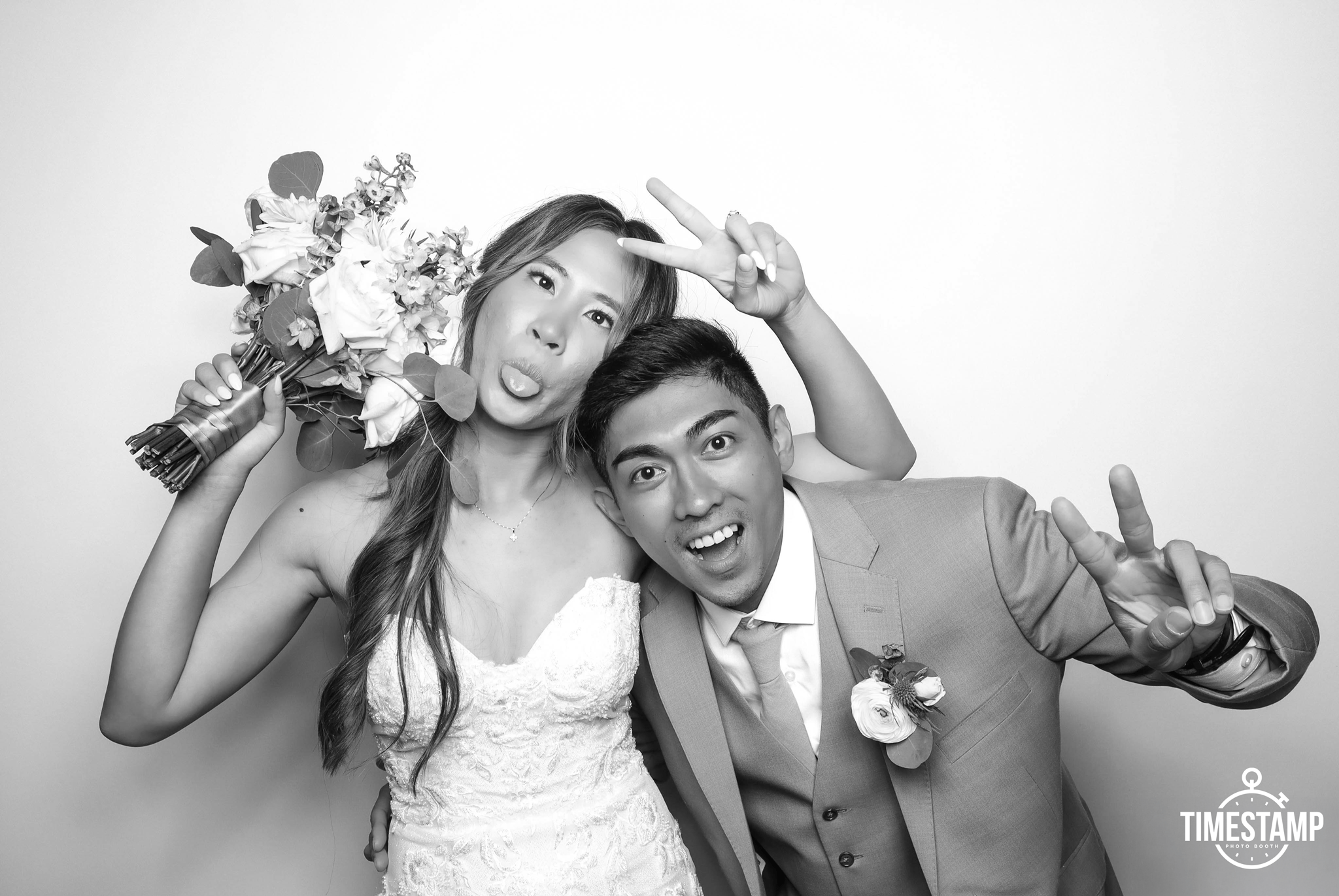 Black and white photo of a joyful bride and groom making funny faces and peace signs at a photo booth, with the bride holding a bouquet of flowers.