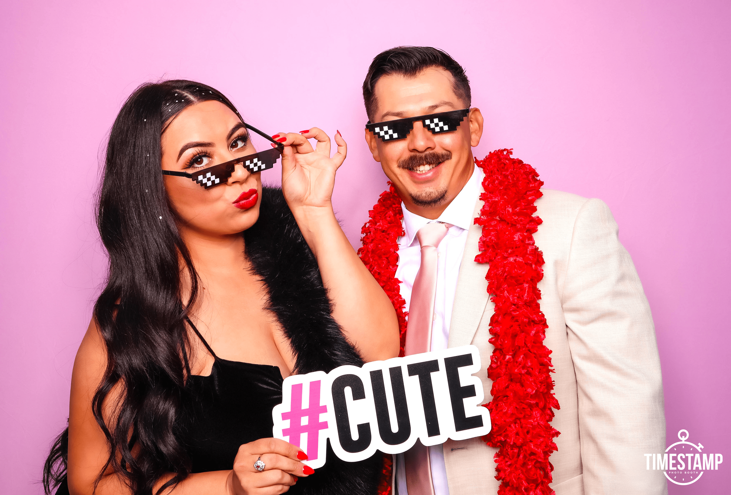 A smiling woman and man wearing pixelated sunglasses, with the woman holding a sign that says #CUTE, standing against a pink background, both dressed in semi-formal attire with fun accessories.