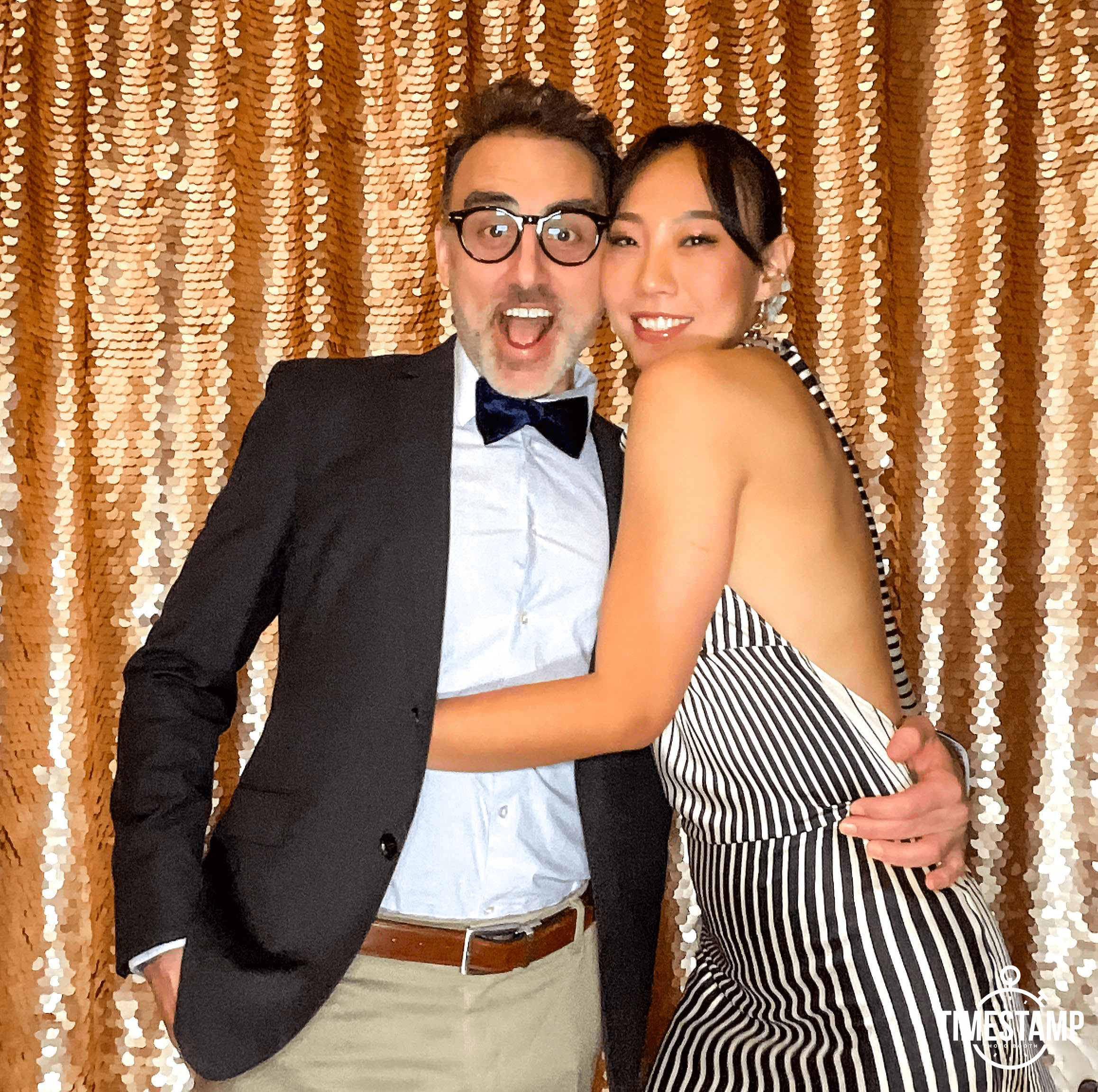 A man and a woman dressed in formal attire, smiling and hugging in front of a gold sequin backdrop.