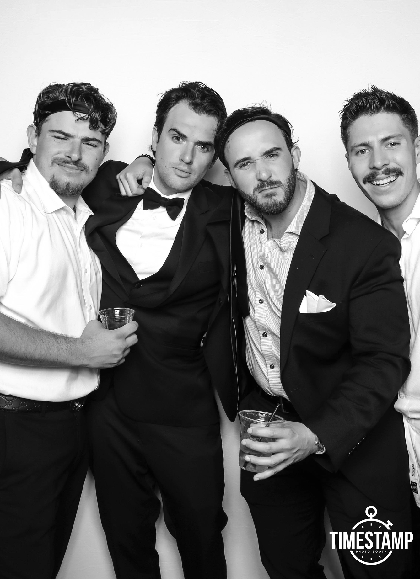 Group of four men dressed in formal attire, holding drinks, posing together at a photo booth, with a Timestamp logo in the bottom right corner.