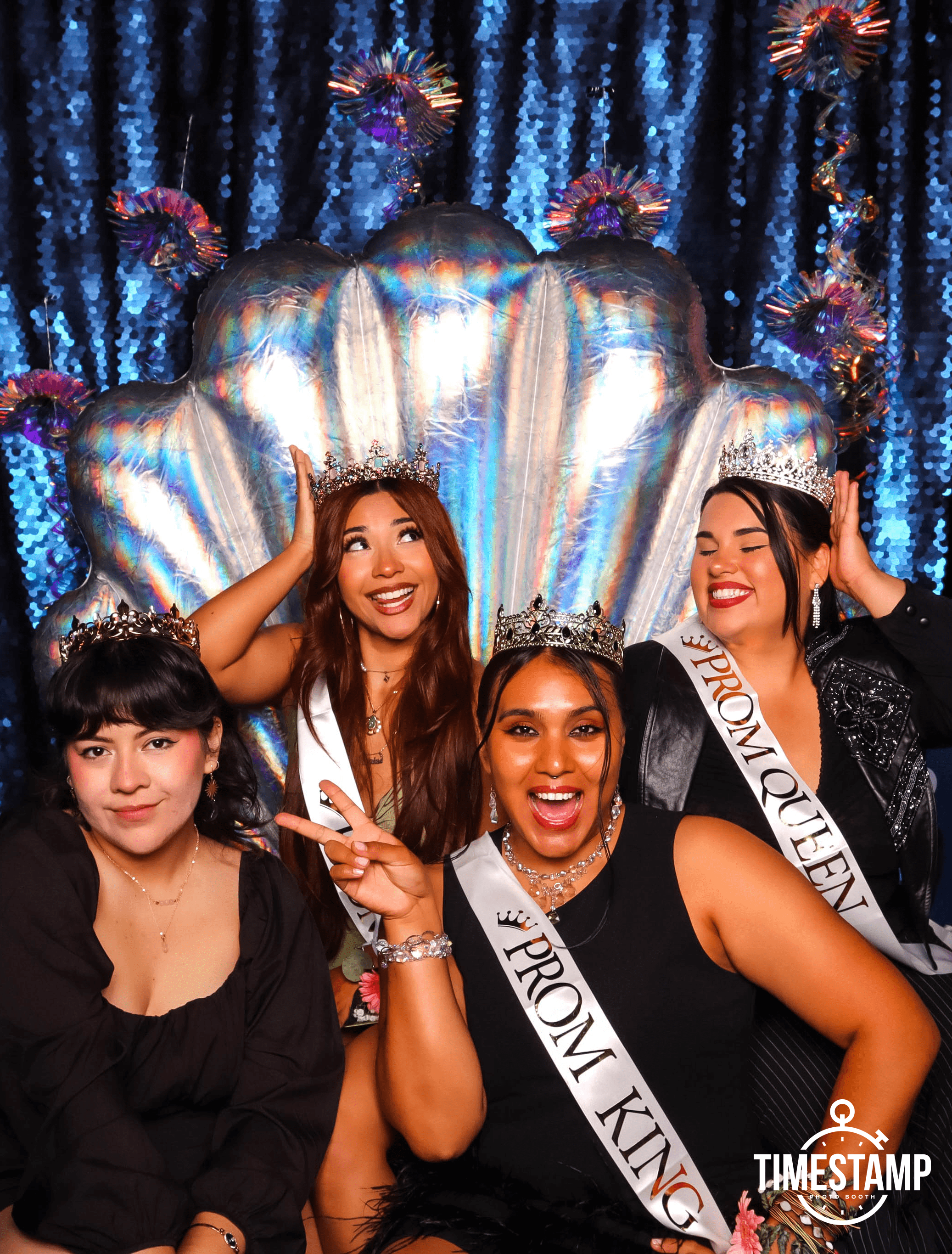 Four women wearing crowns and sashes that say 'Prom Queen' and 'Prom King' in a photo booth with a blue sequin backdrop and a large iridescent shell decoration, having fun at prom.