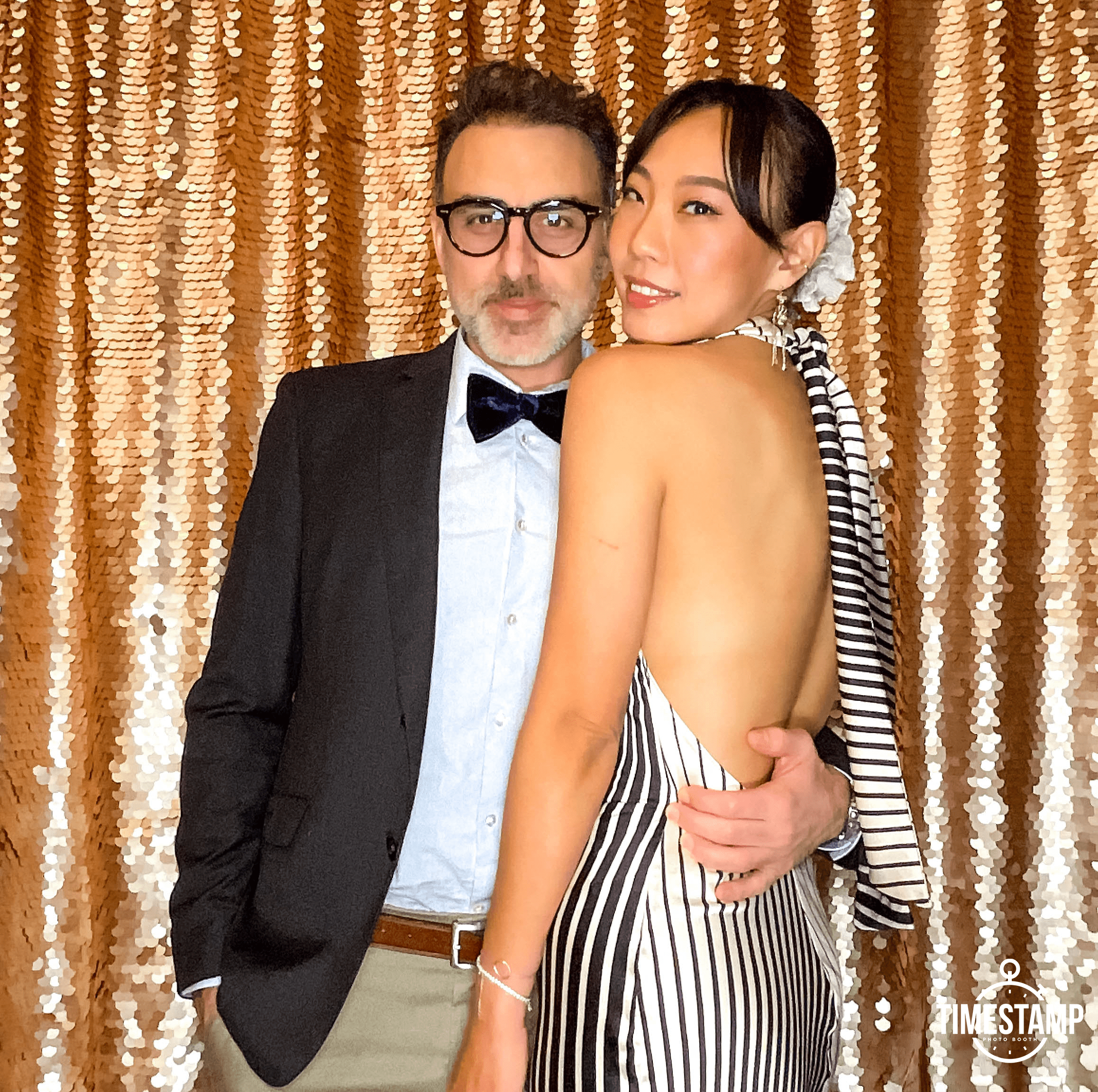 A man in a suit with glasses and a woman in a striped dress with a white flower in her hair, posing together in front of a sparkling gold sequin backdrop.