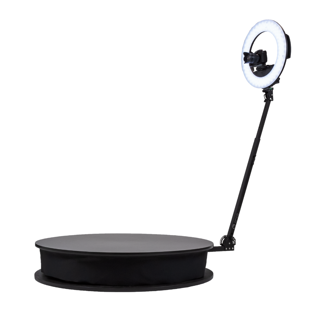 A ring light with a camera mounted in the center, attached to an adjustable stand, positioned facing a round platform with a soft surface.
