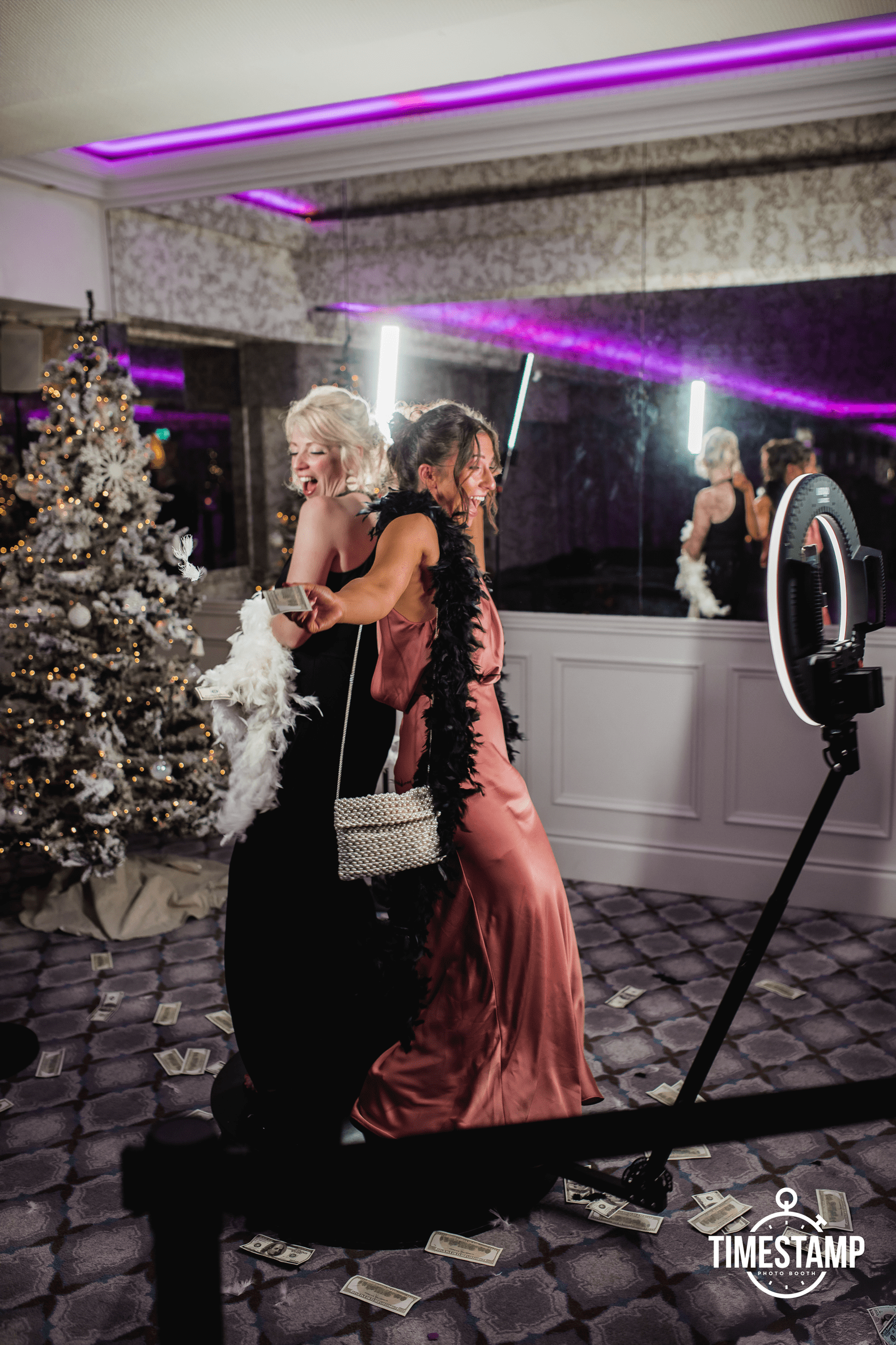 Two women dancing and celebrating at a holiday party with a decorated Christmas tree, confetti, and dollar bills on the floor, illuminated by purple and pink lighting.