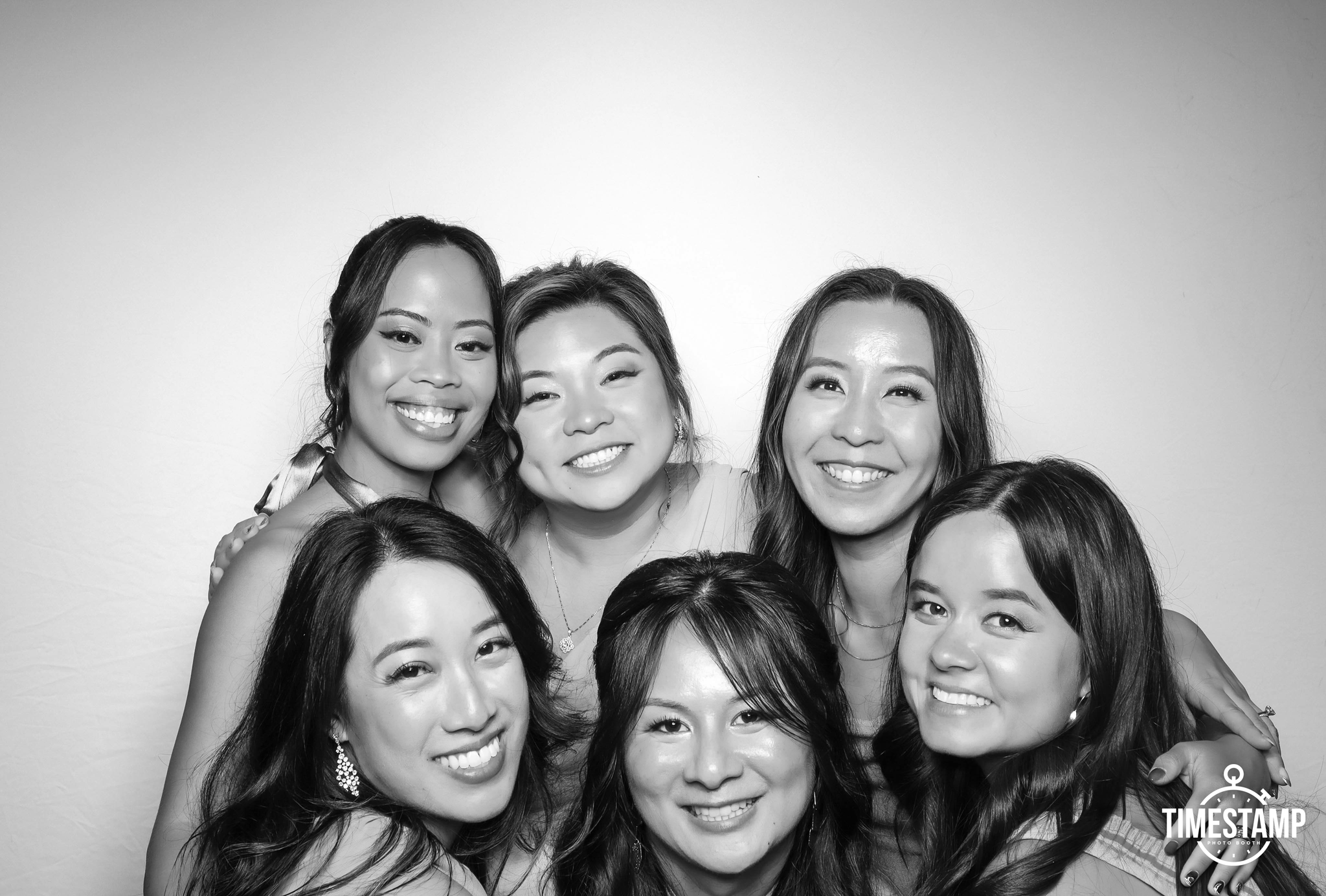 Group of seven women smiling and posing together in a photo booth with a plain background, black and white photography.