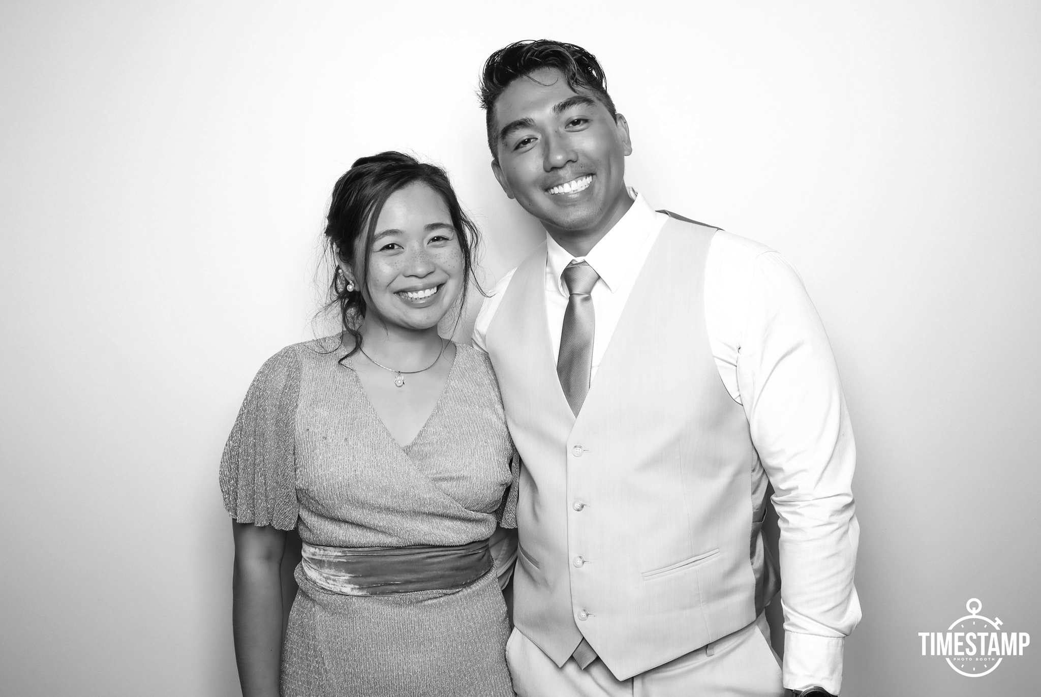 A smiling couple dressed in formal attire standing together against a plain background, with the logo "TIMESTAMP" in the bottom right corner.
