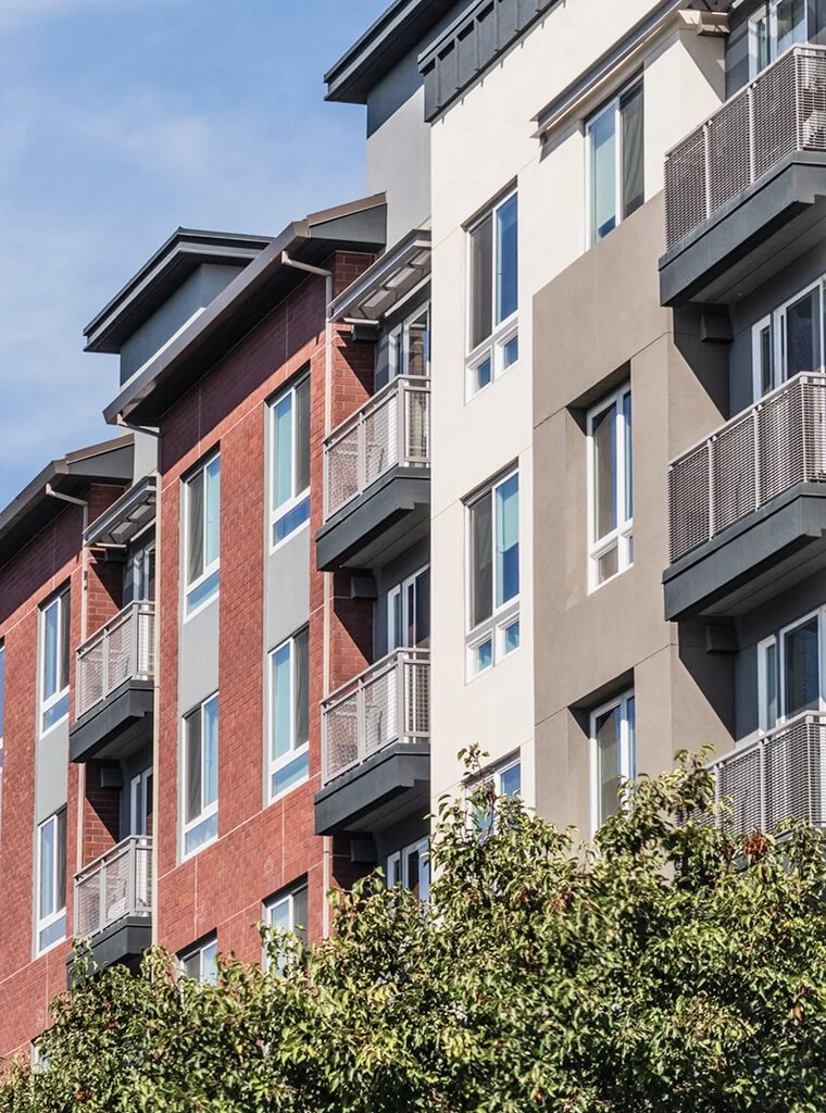 A modern multi-story apartment building with red brick and beige stucco exterior, featuring multiple balconies and windows, with a tree in the foreground under a blue sky.