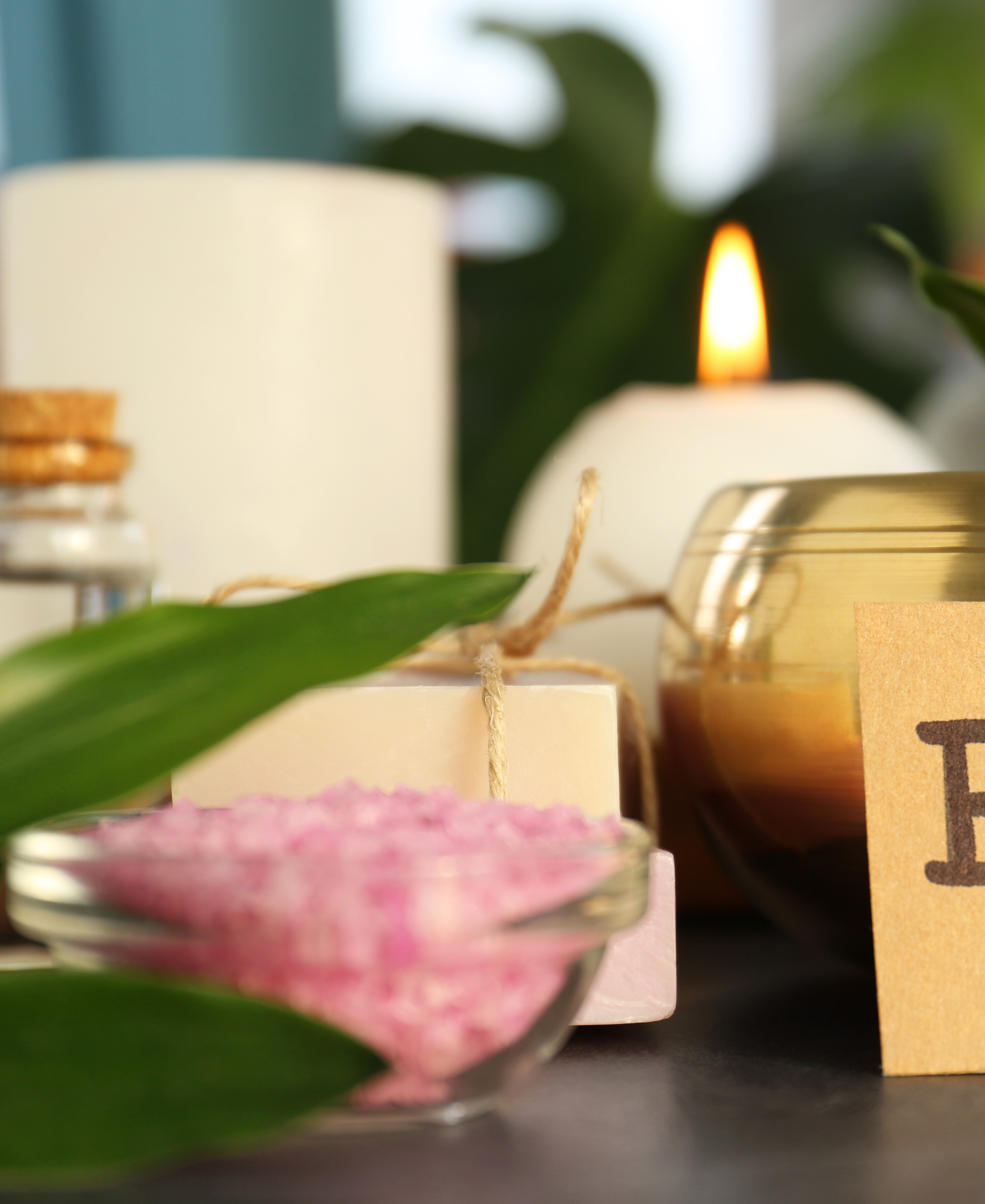 Various candles, a pink bath salt in a glass bowl, green leaves, and a small card on a table, creating a relaxing spa or wellness setting.