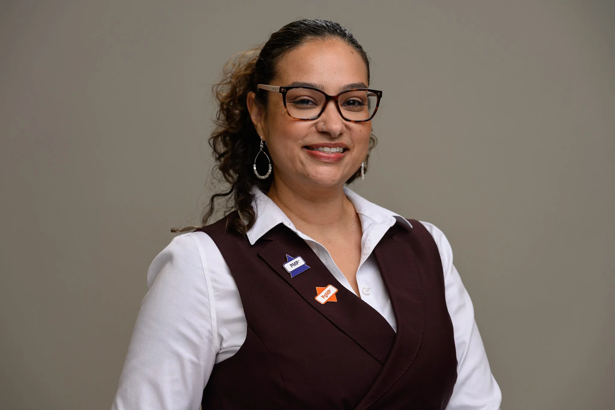A woman with glasses and earrings, smiling, wearing a white shirt and maroon vest with pins, standing against a neutral background.